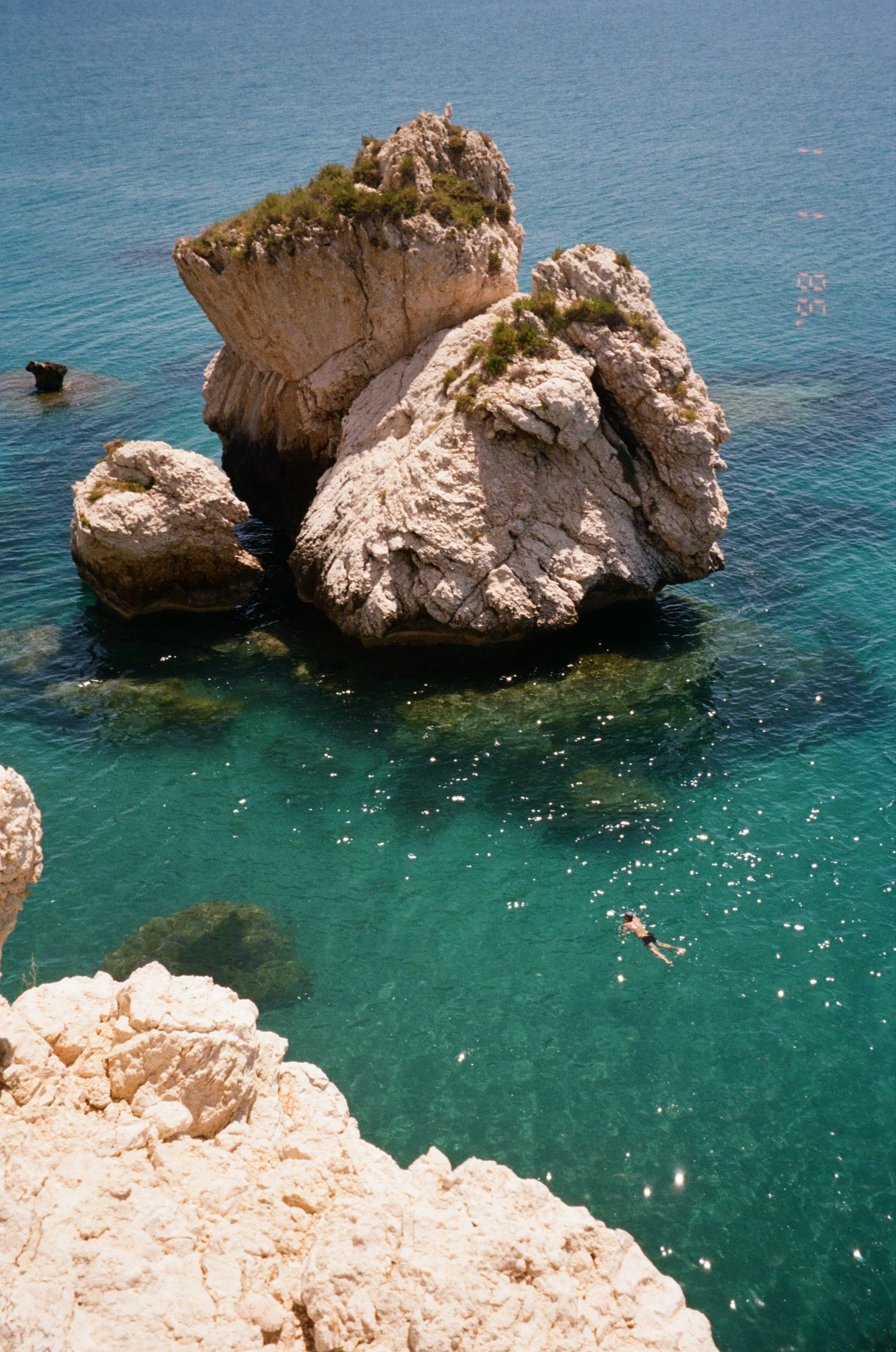 Large rocky formation in turquoise water with a person swimming nearby, and rocky cliffs in the foreground.