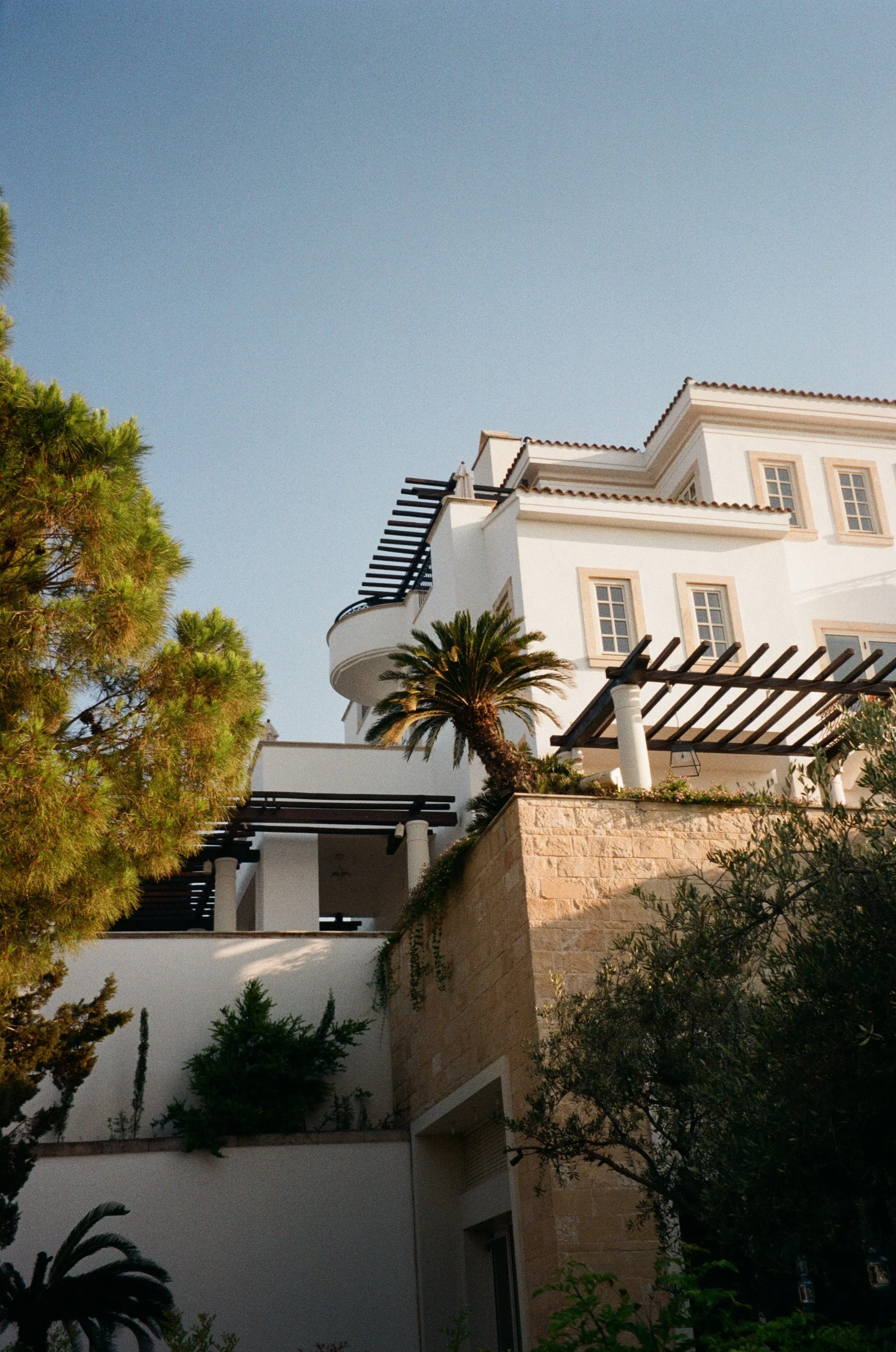 A multi-story white Mediterranean-style house with beige trim, outdoor balconies with black pergolas, and a rooftop terrace. There are palm trees and greenery in the foreground under a clear sky.