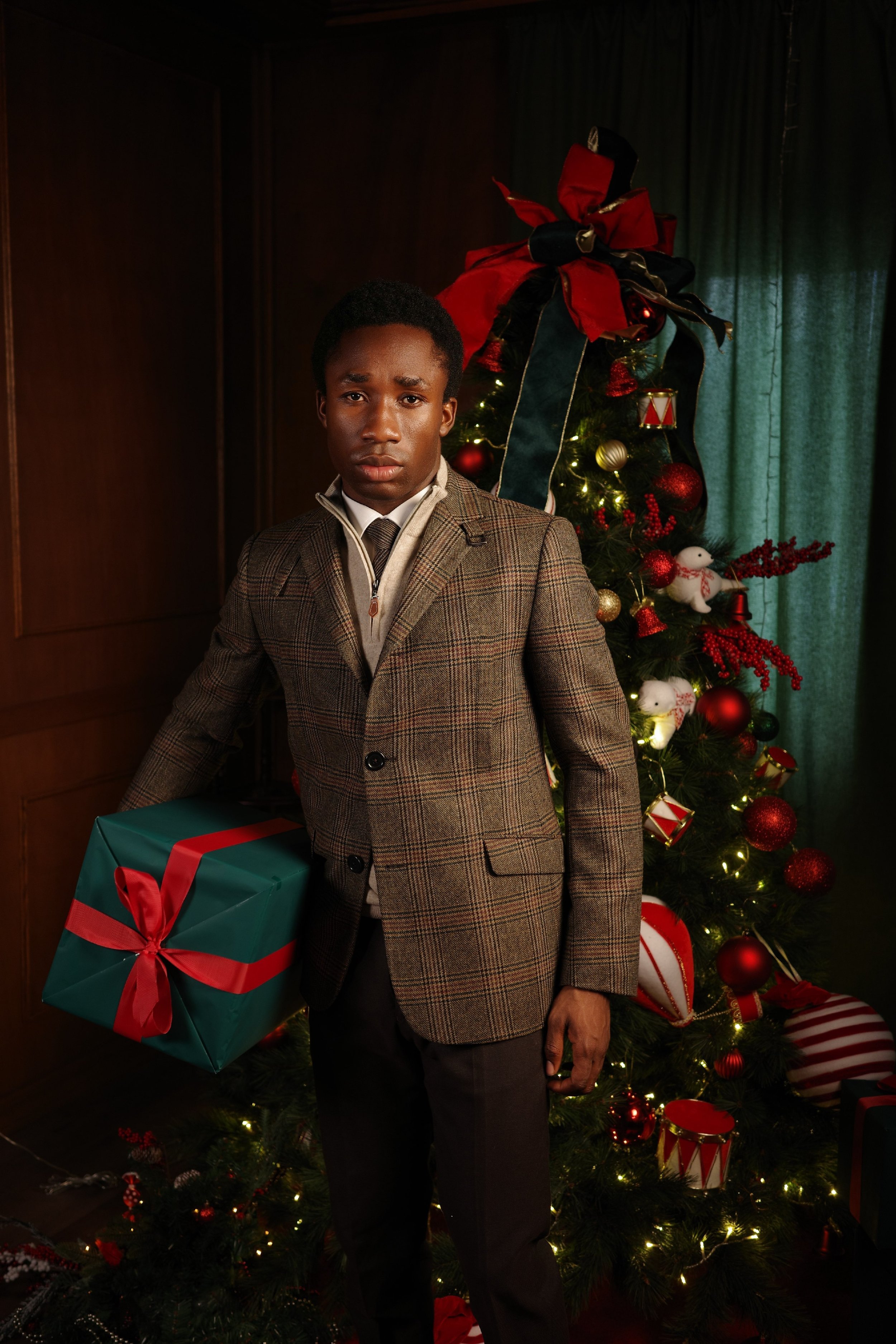 A young man dressed in vintage attire holding a wrapped Christmas gift, standing in front of a decorated Christmas tree with lights and ornaments.