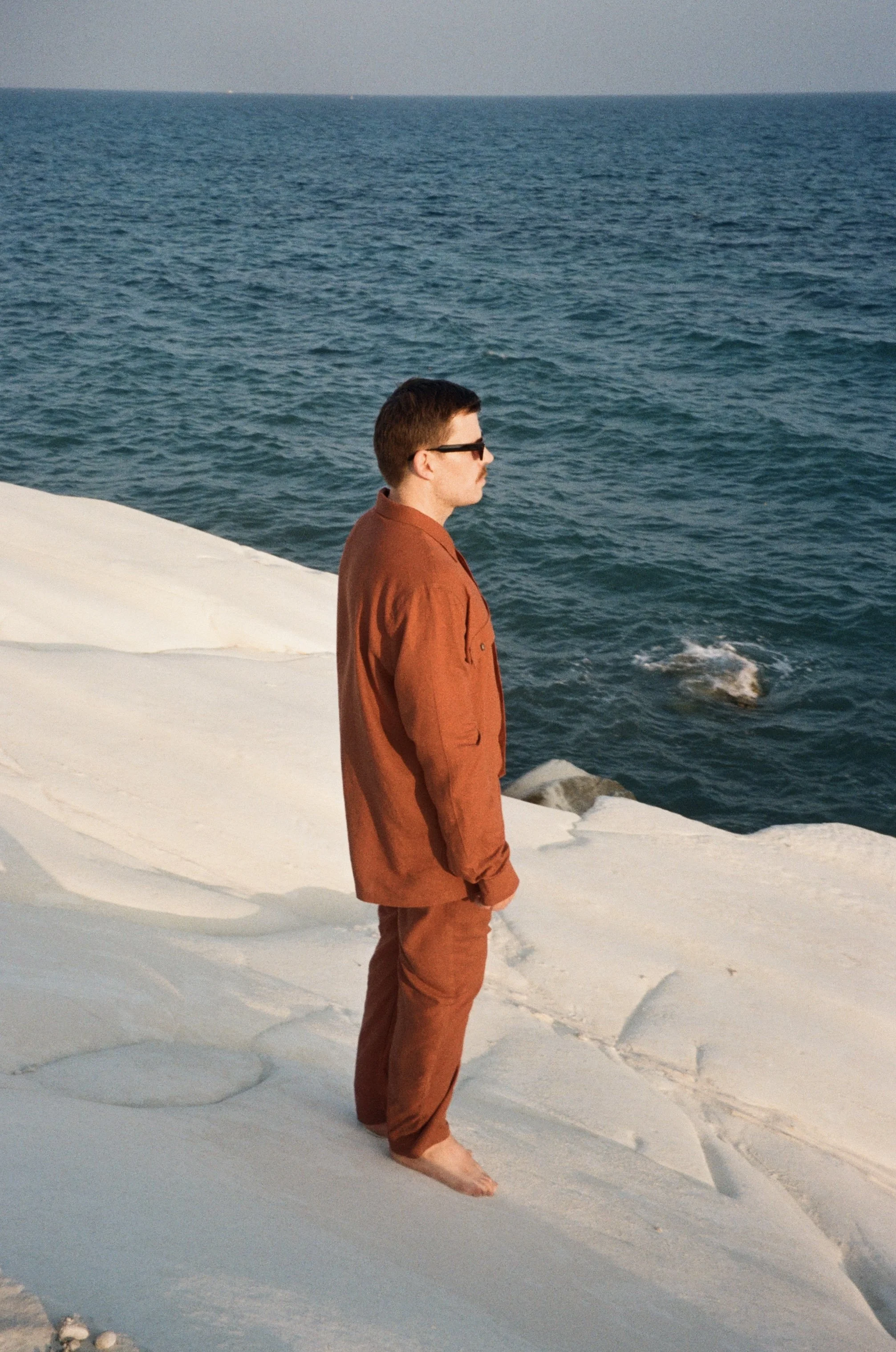 A man in a brown jacket and pants, barefoot and wearing sunglasses, standing on white rocky terrain by the ocean, looking out at the water.