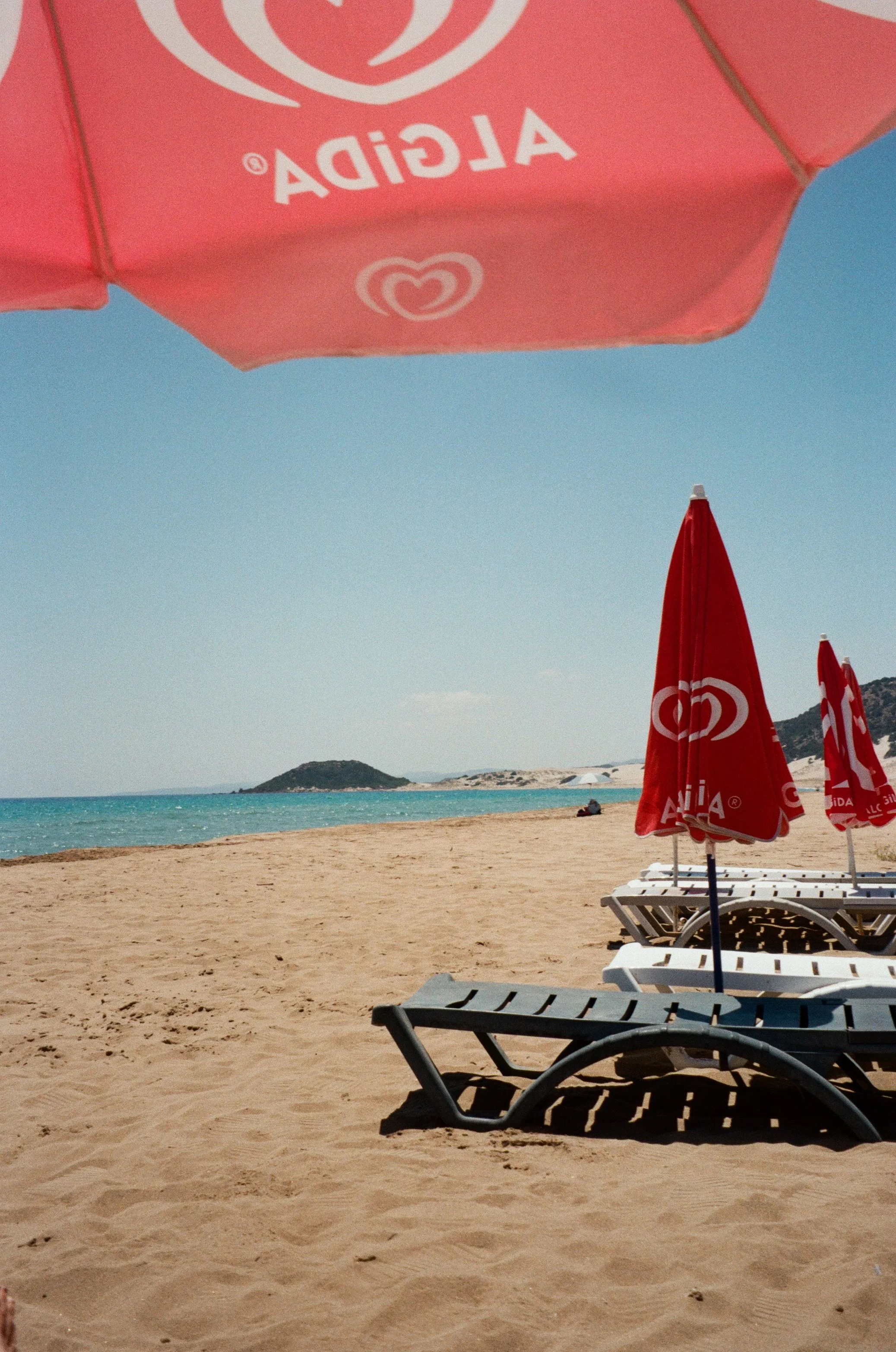 Beach scene with sand, blue ocean, small island in the distance, multiple red and white umbrellas with ice cream brand logo, and lounge chairs on the sandy beach.