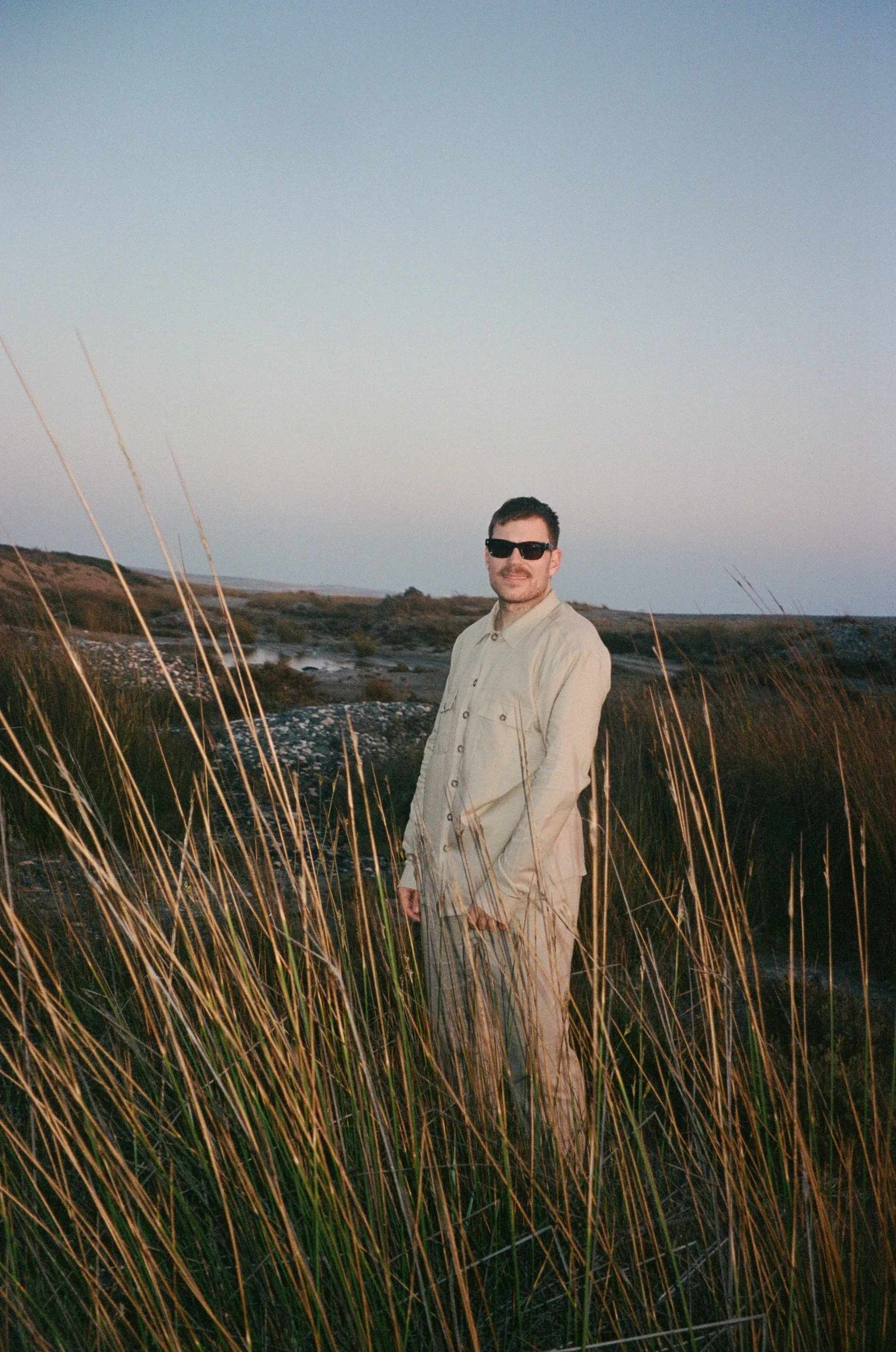 A man wearing sunglasses and light-colored clothing standing among tall grass in a scenic outdoor landscape at sunset.