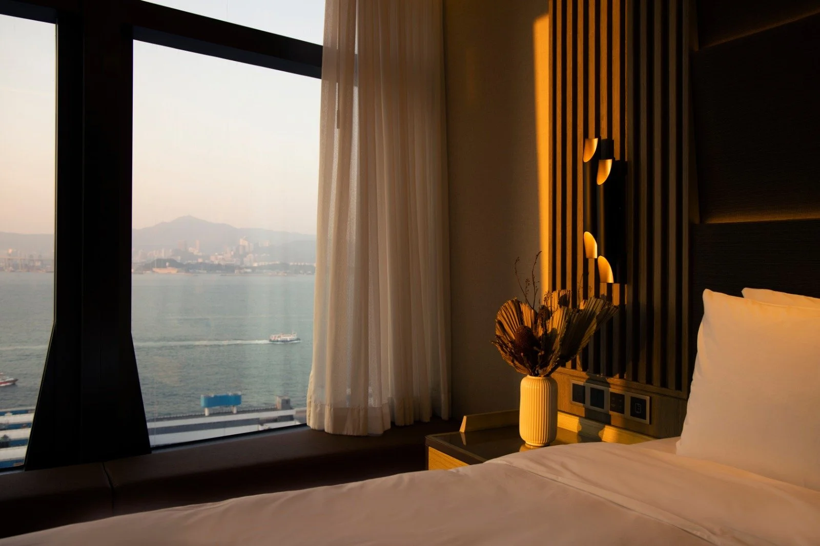 Hotel room with a large window showing a city skyline and water with boats, a bedside table with a vase of dried flowers, and a bed with white sheets.