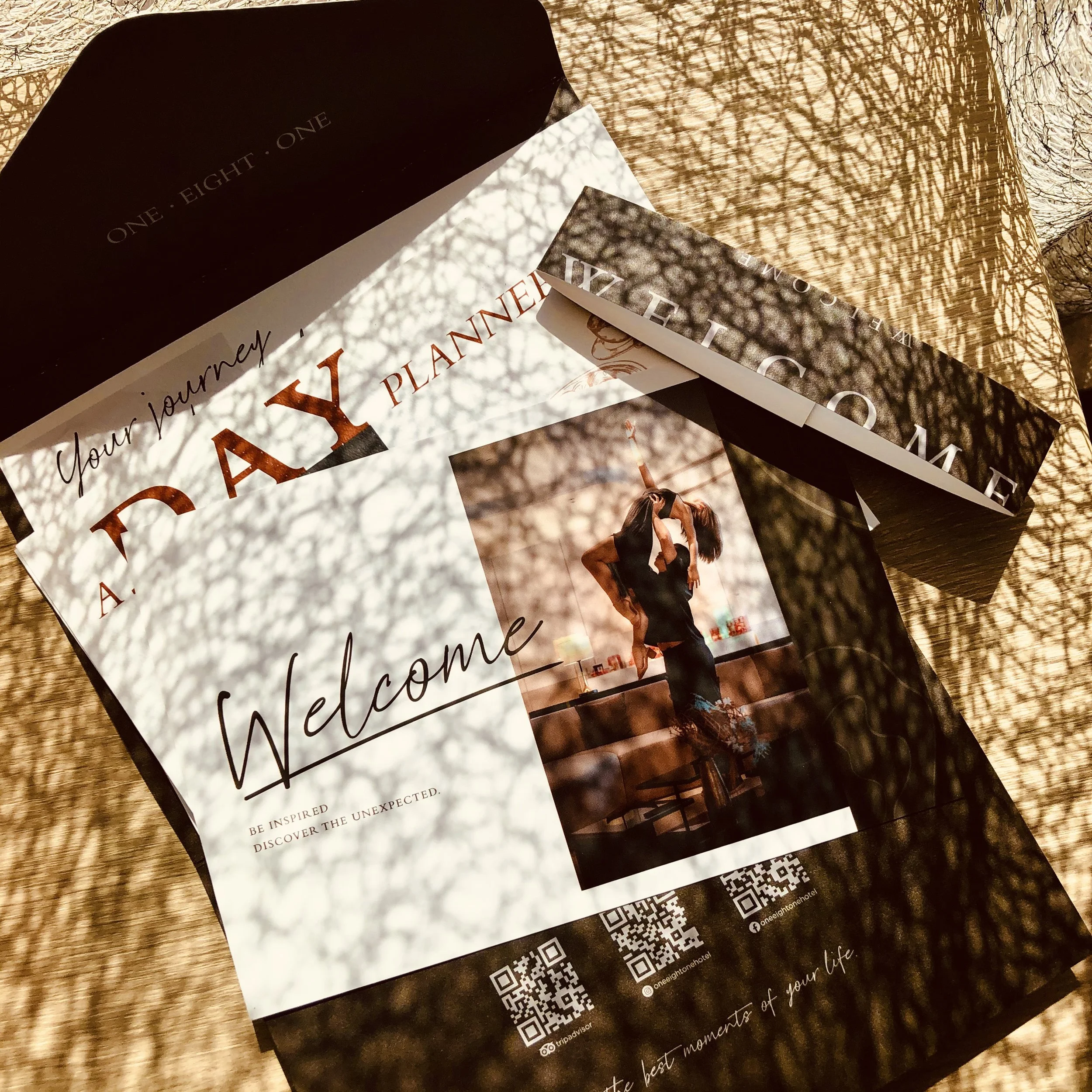 A flat lay photo of a welcome brochure for a travel planner titled 'Your journey A.TAXY PLANNER'. The brochure features a woman doing a yoga pose in a modern indoor setting. The sunlight creates intricate shadows on the surface, and there are QR code