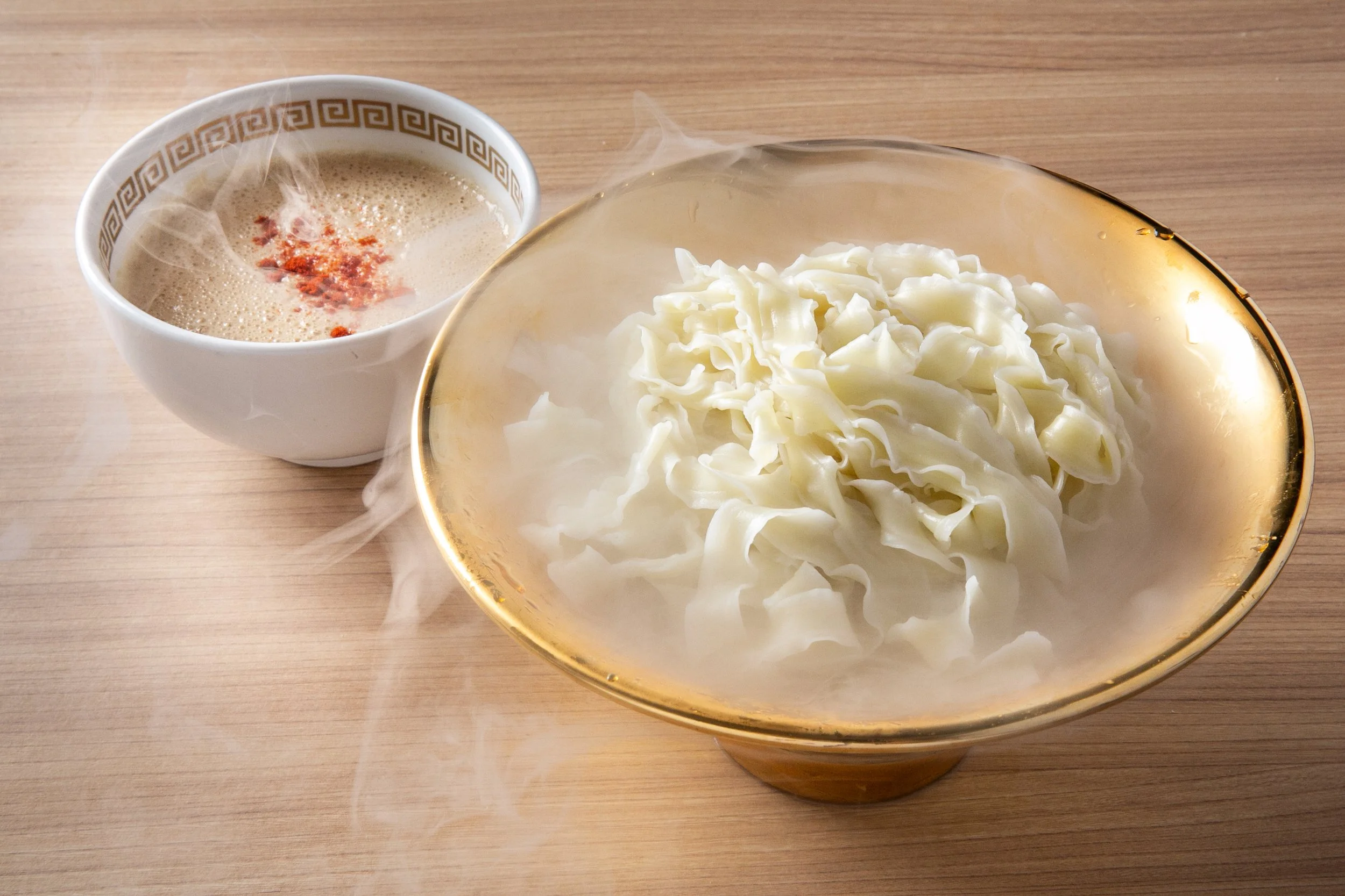 A bowl of steaming hot shredded cabbage with a bowl of soup garnished with red seasoning on a wooden surface.