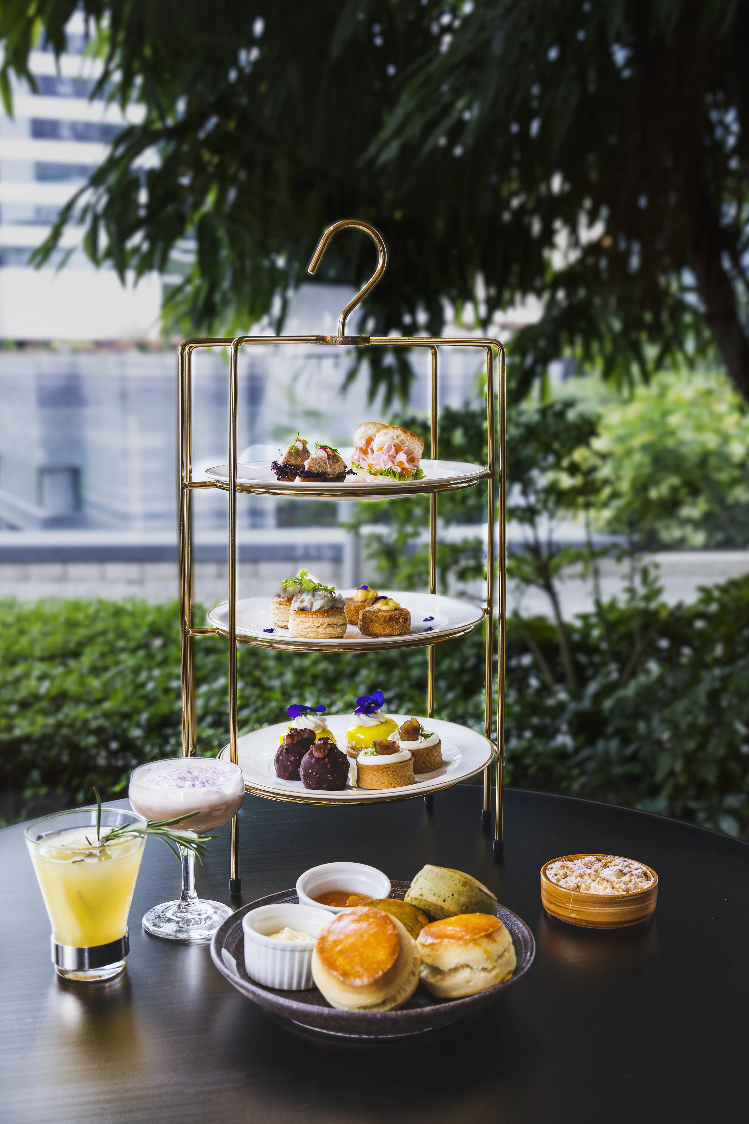 A tiered serving tray with assorted desserts and beverages on a black table outdoors, with greenery and city buildings in the background.