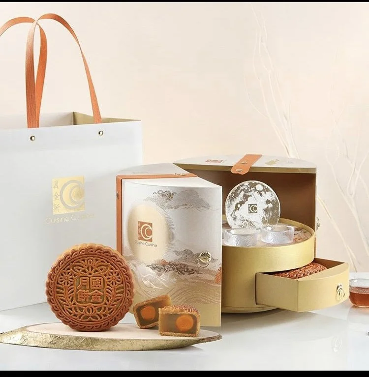 Assorted mooncakes in elegant packaging, including a whole mooncake with intricate design and a slice showing filling and yolk, displayed on a reflective surface with a pastel background.