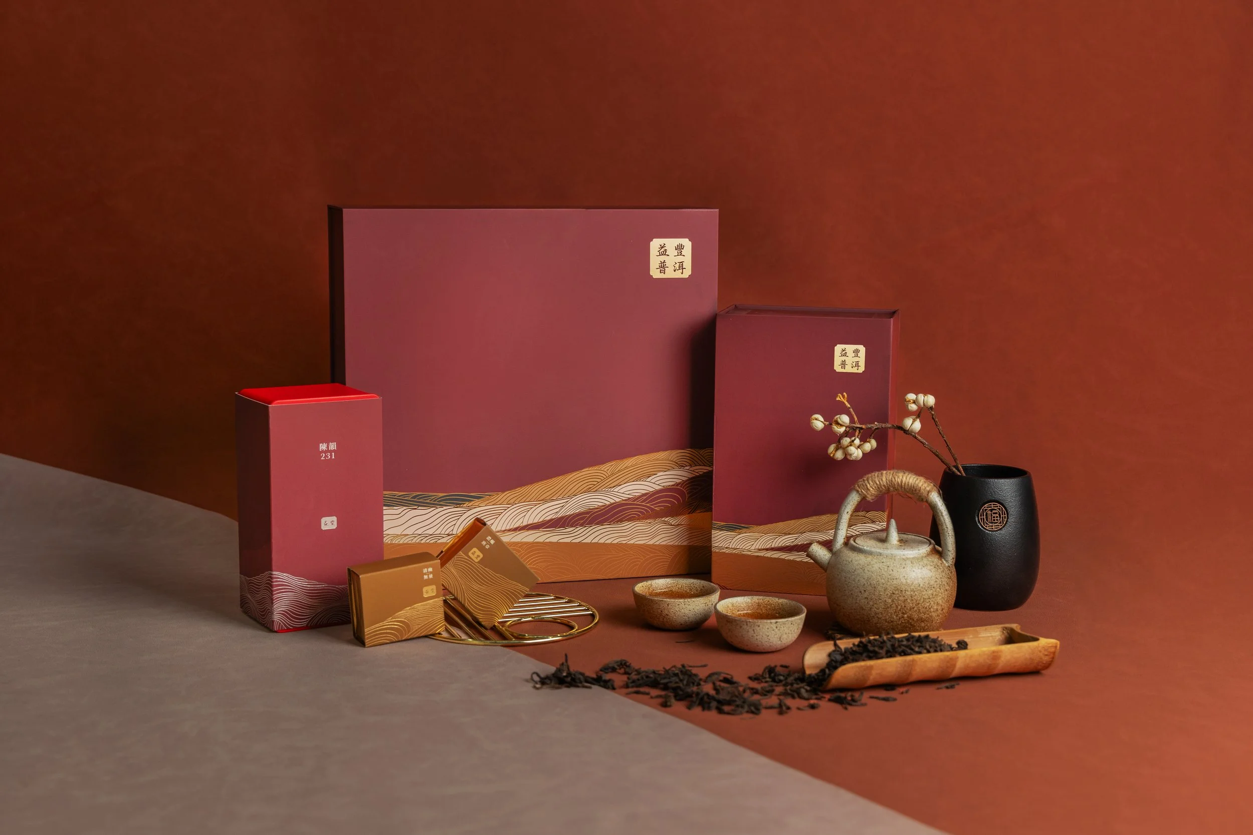 Tea set with a teapot, two cups, and loose tea leaves, surrounded by pink and gold boxes, a black vase with flowering branch, set against a brown and beige background.