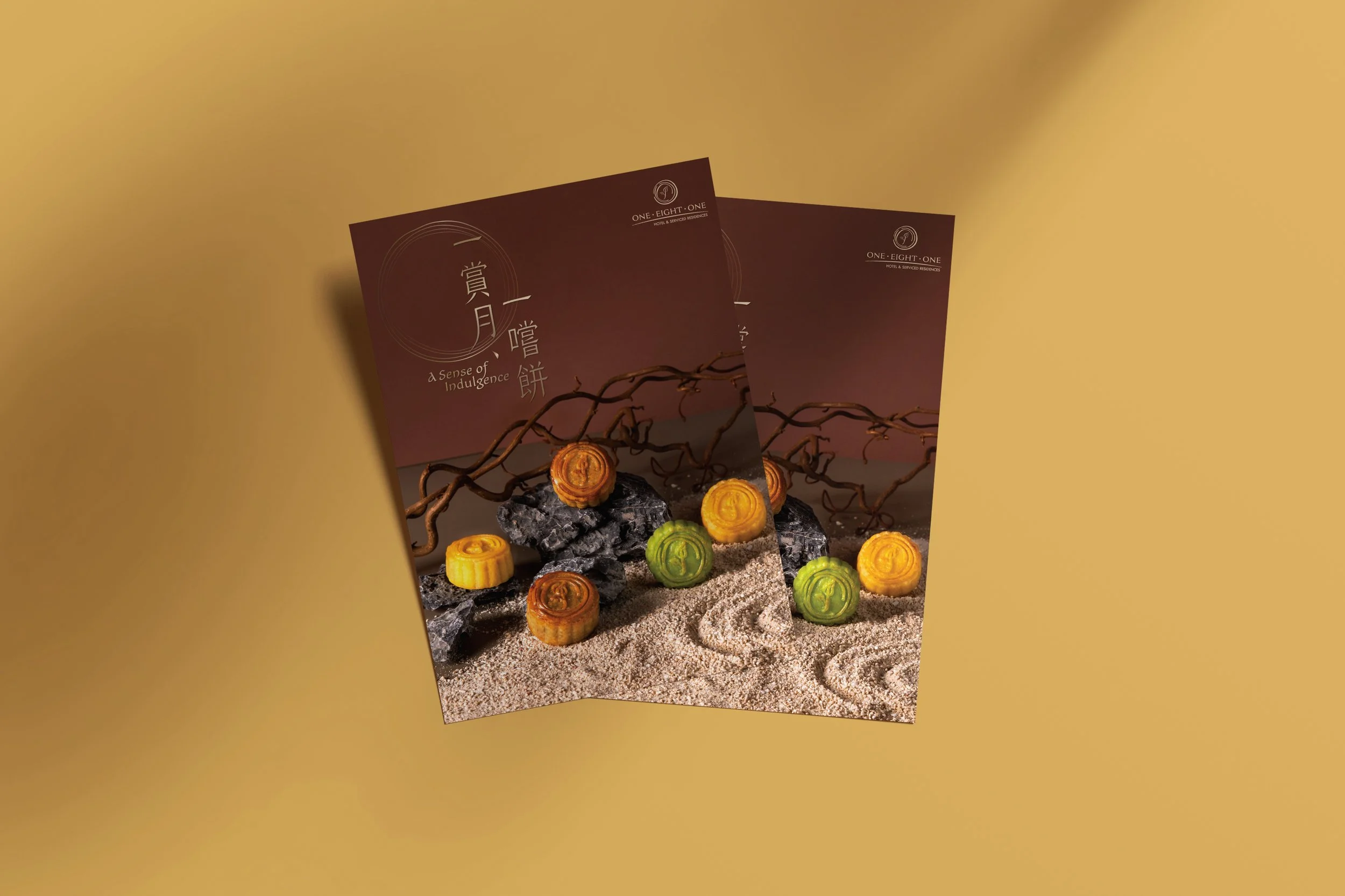 Two brochures with a scenic design of rocks, sand, and branches, displaying colorful mooncakes, branded as 'A Sense of Indulgence,' from One Eight One hotel or resort.