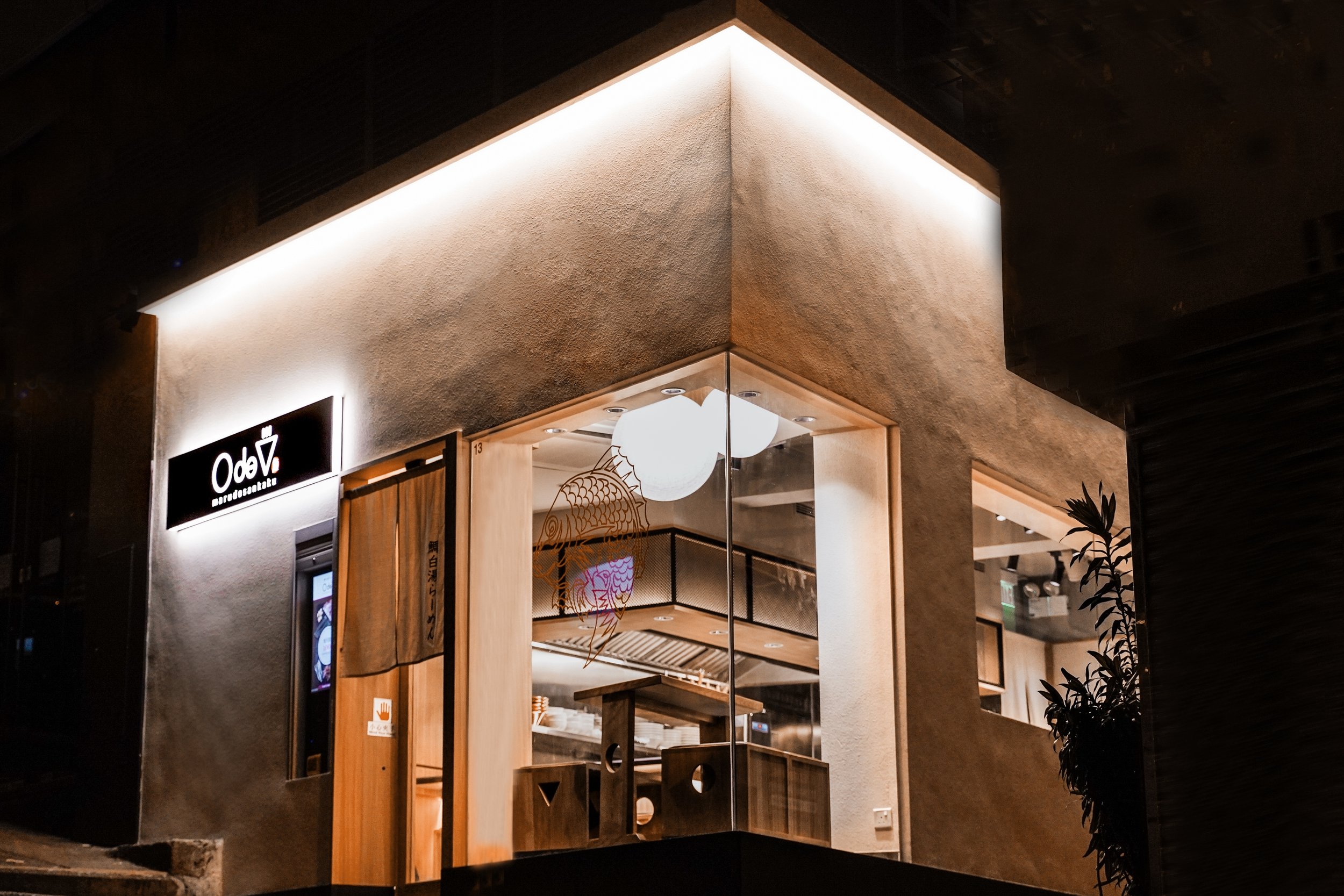 Night view of a modern restaurant with illuminated signage and large glass windows, showing a cozy interior with lighting fixtures and wooden furnishings.