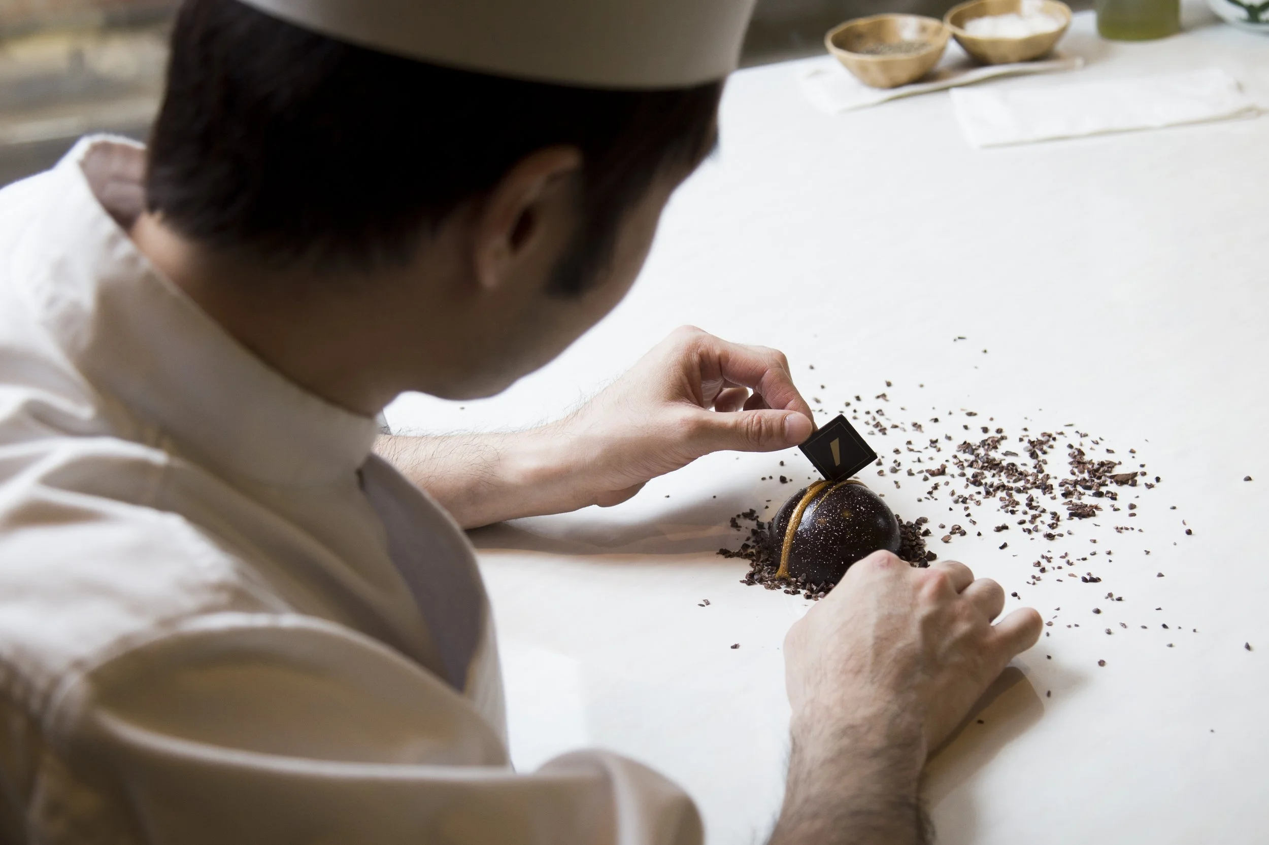 A chef decorating a spherical chocolate dessert with a small tool on a white workspace, with bowls of ingredients in the background.