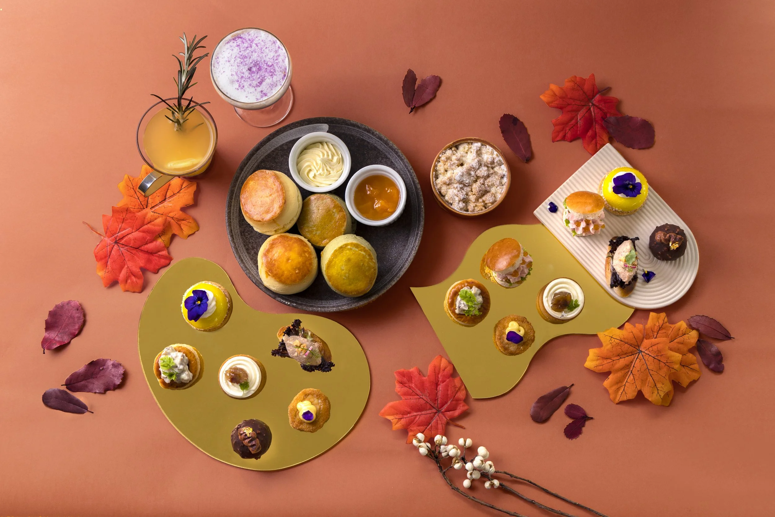 Autumn-themed spread of small pastries and desserts on decorative platters, surrounded by colorful fall leaves.
