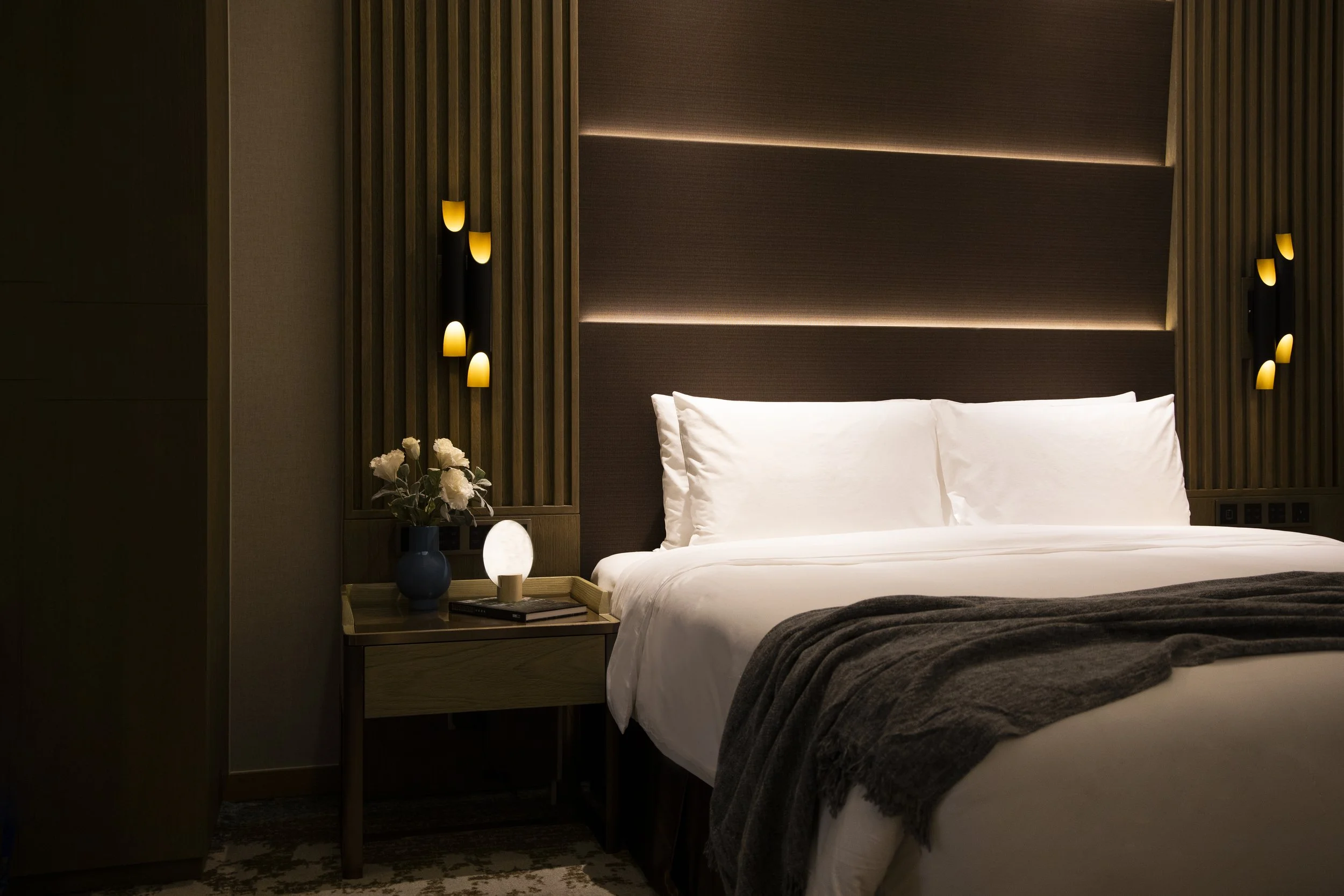 A bedroom with a neatly made bed featuring white pillows and sheets, a dark throw blanket, a wooden side table with a vase of flowers and a lamp, and wall-mounted light fixtures on a padded headboard wall with vertical wooden slats.
