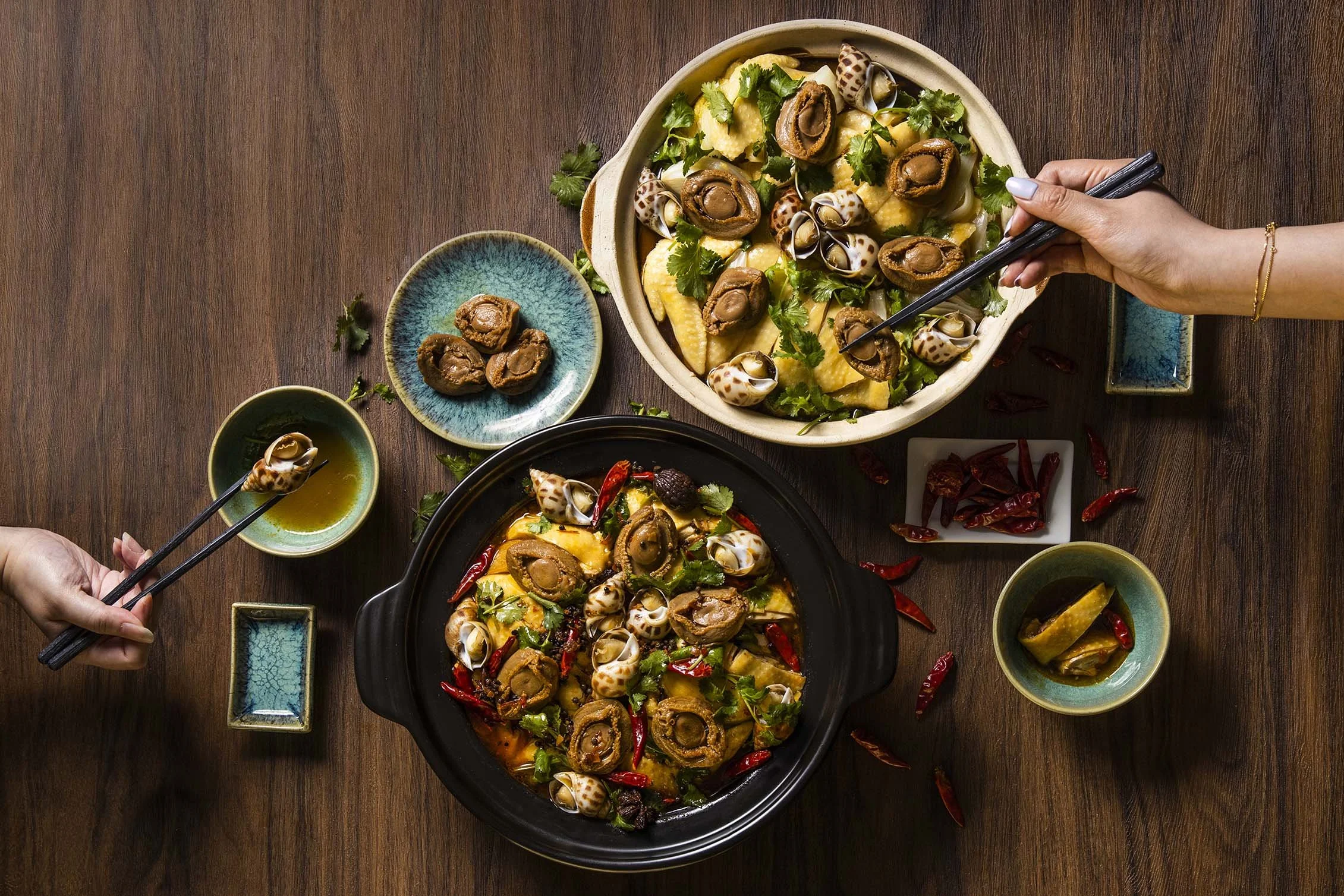 Two people sharing a meal of spicy seafood and vegetables, including snails, peppers, and herbs, in black and white bowls on a wooden table.