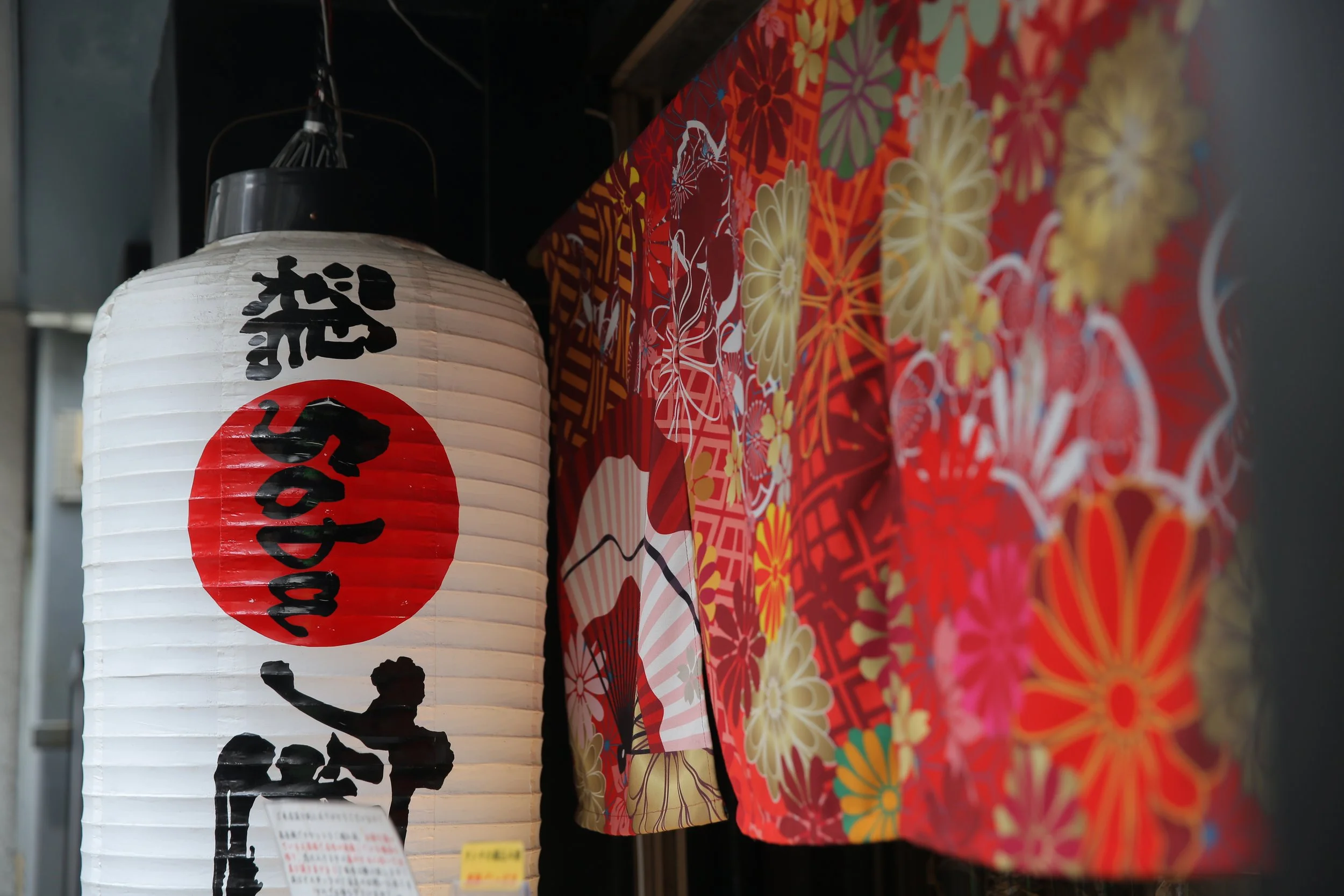 A white Japanese paper lantern with black and red Japanese characters and a silhouette of a person, hanging next to colorful floral patterned fabric.