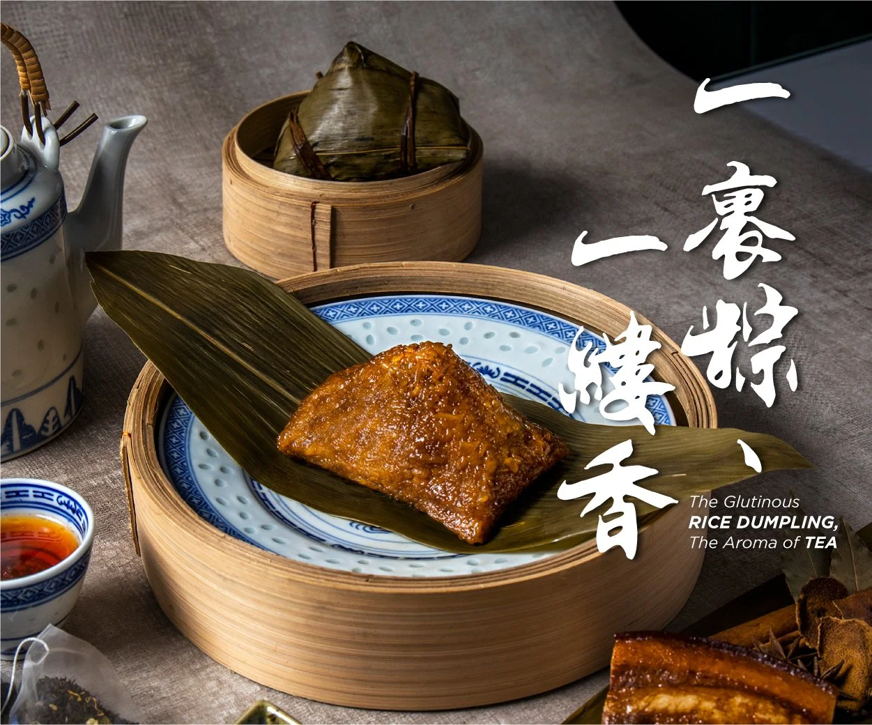 A traditional Asian rice dumpling wrapped in bamboo leaves, placed on a decorative blue and white plate inside a round bamboo steamer. Surrounding items include a small bowl of sauce, a teapot, and additional wrapped items.
