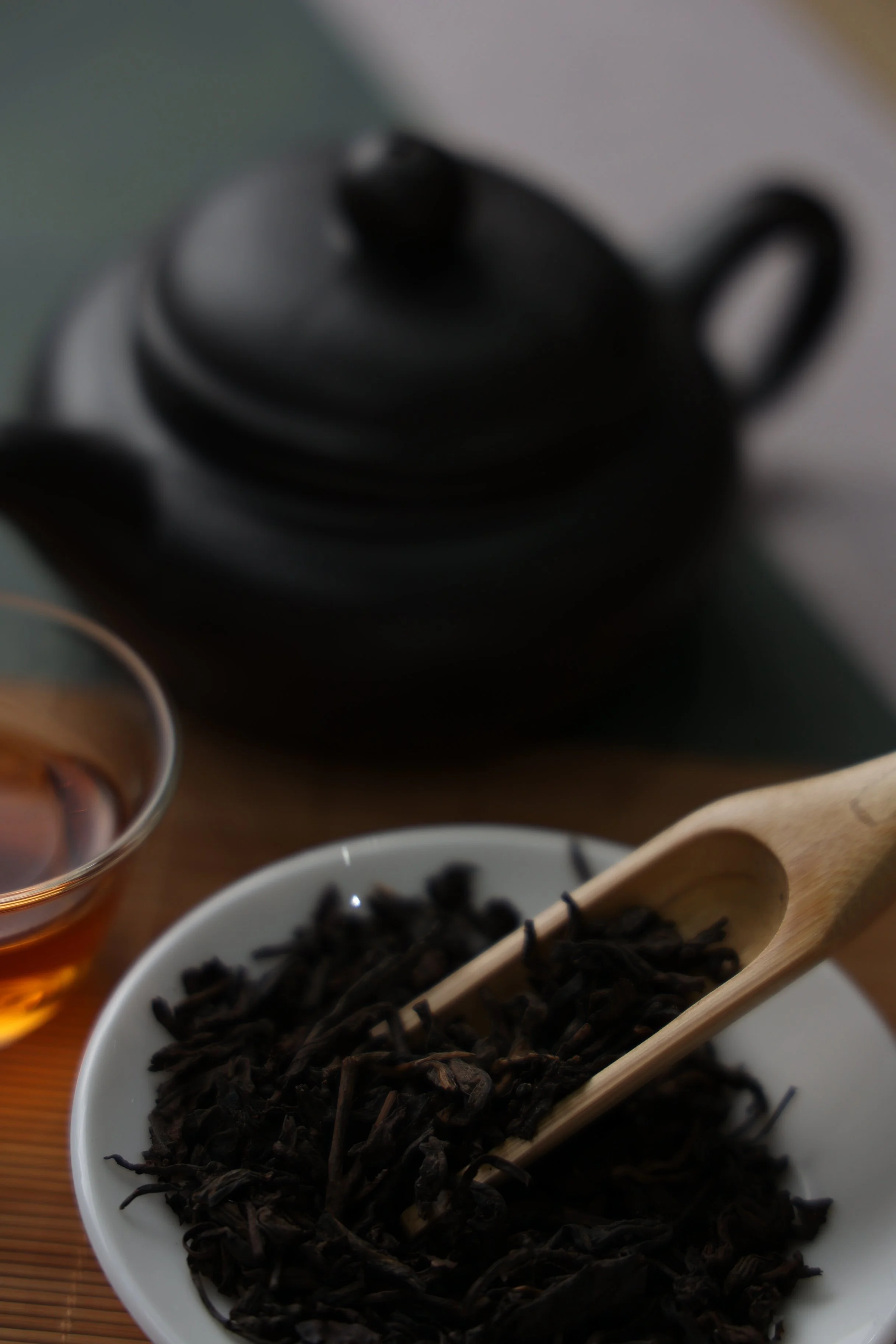 Close-up of a bowl of dried black tea leaves with a wooden scoop, a glass cup of brewed tea, and a black teapot in the background.