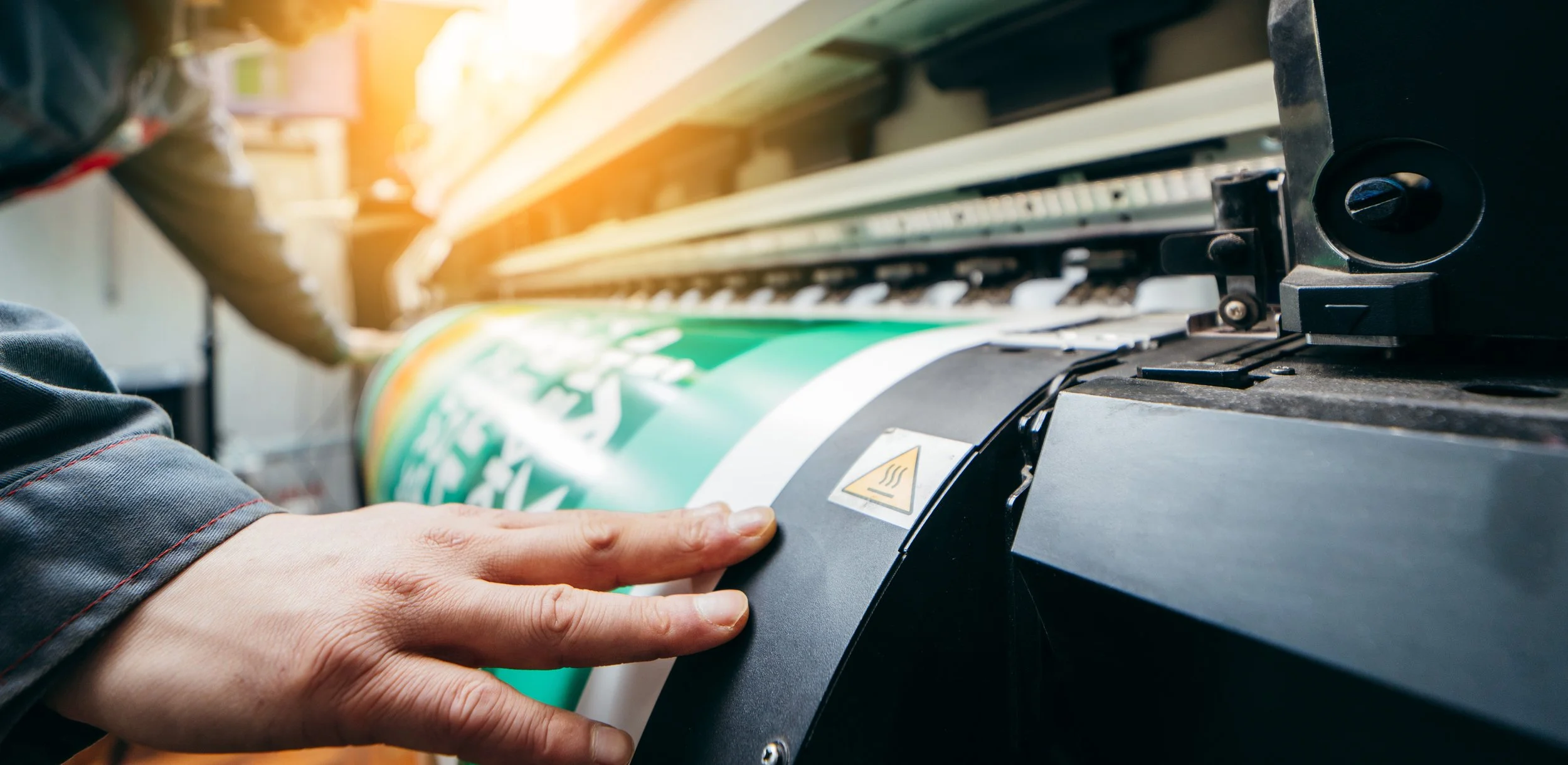 A person's hand printing on a sheet using a large-format printer, with a focus on the printer's components and a warning label.
