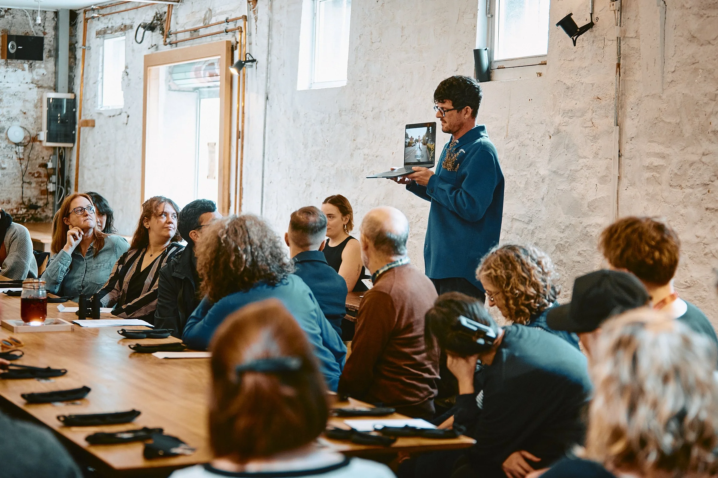 A man in a blue shirt stands in front of a group of people sitting at a long wooden table in a rustic room with white brick walls, holding a laptop. The attendees are listening and some are taking notes.