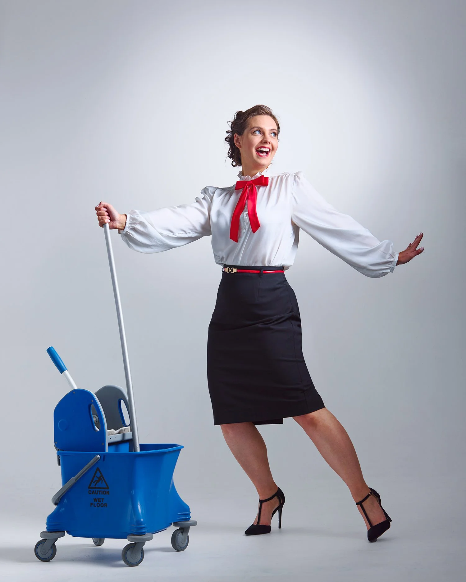 Woman smiling, dressed in office attire with a red ribbon, posing next to a blue mop bucket and cleaning tools in a studio setting.