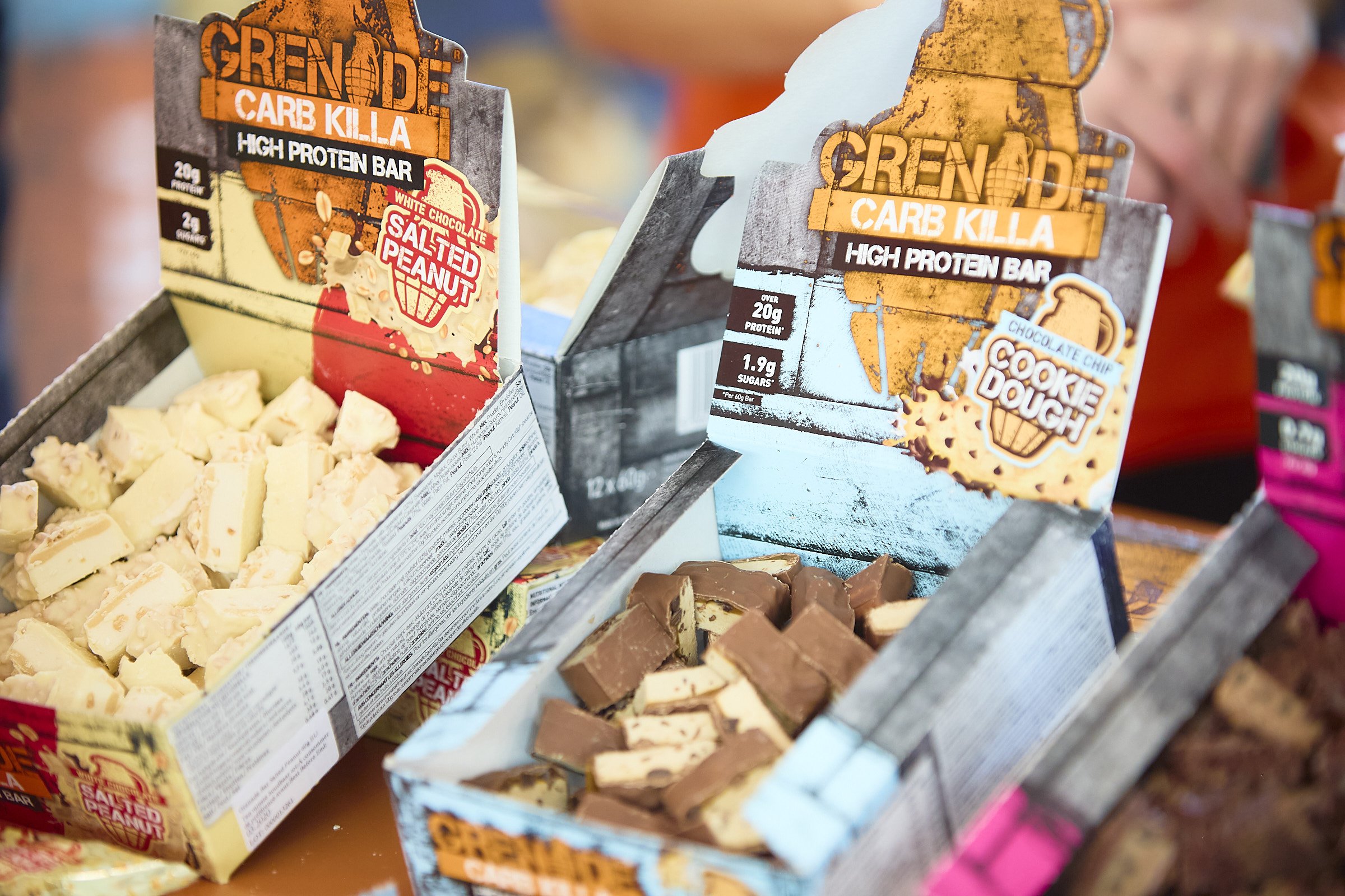 Assorted Greneade Carb Killa protein bars in brightly colored packaging, including Salted Peanut and Cookie Dough flavors, displayed in open cardboard boxes.