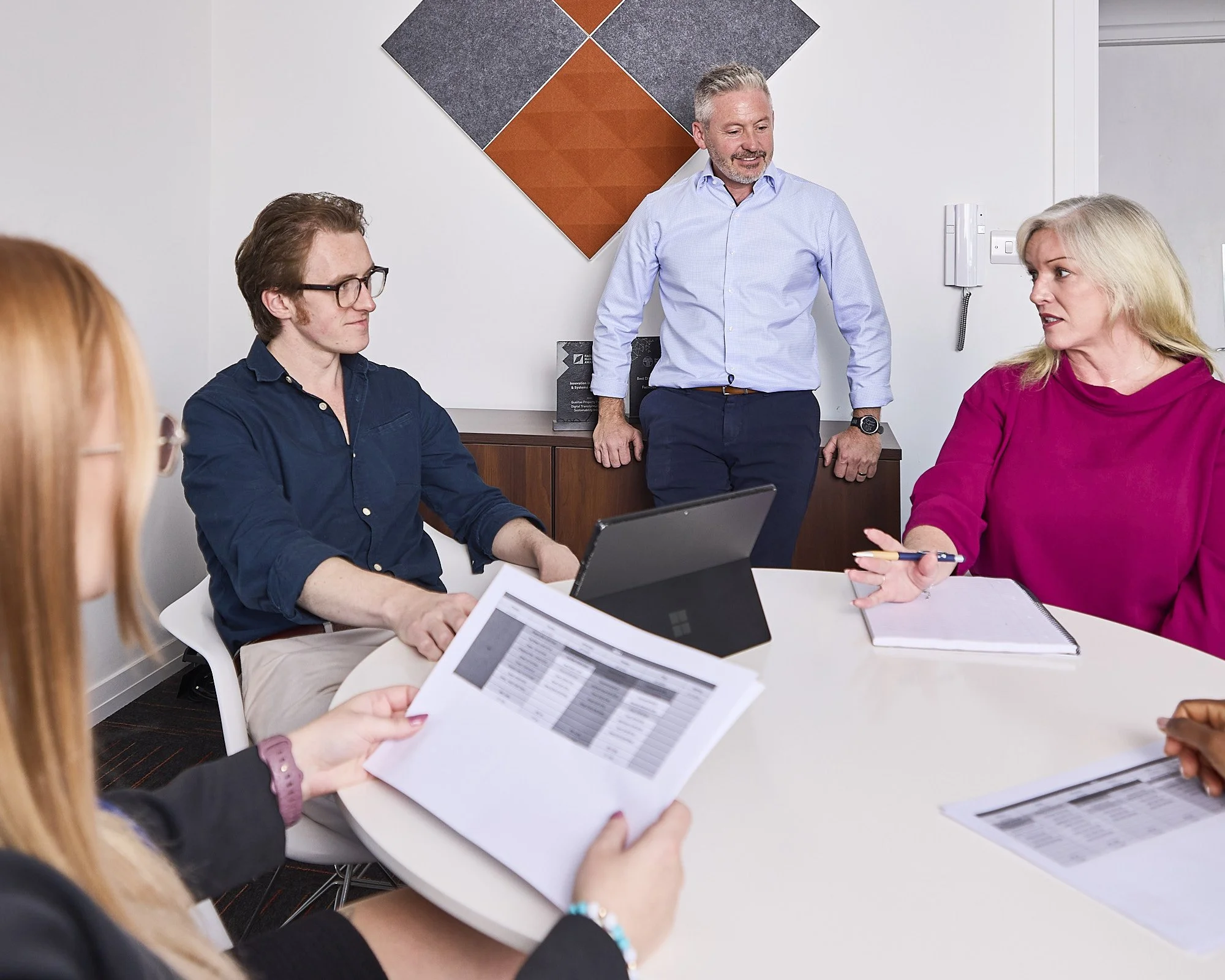 Professional photo of a group of five professionals in a meeting room, with one woman speaking and others listening, with documents, a laptop, and a tablet on the table. location photography. 