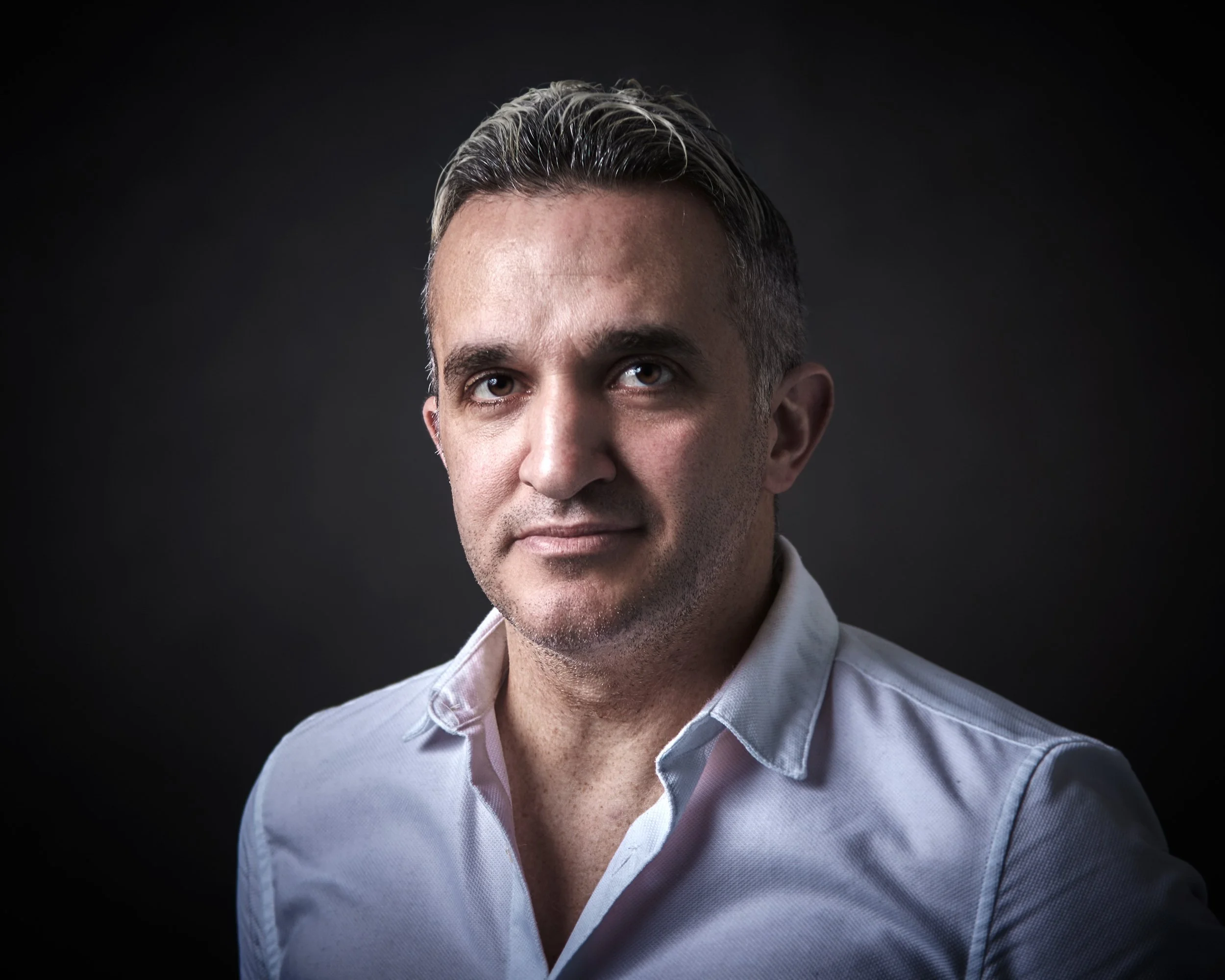 A professional headshot of a man with short, dark, and gray hair wearing a white shirt against a dark background in a professional photo studio with high quality lighting .