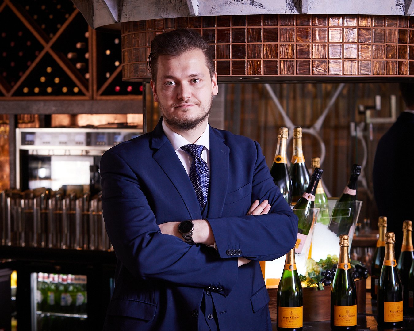 A man in a navy blue suit and tie with arms crossed, standing in a wine bar or restaurant, surrounded by bottles of sparkling wine and wine glasses, with a wooden wine rack in the background.