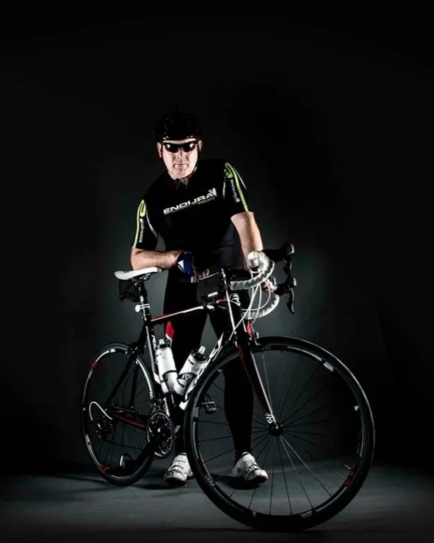 Advertising profession photo taken in studio of a male cyclist in black and green cycling gear, wearing sunglasses, posing with his black and red road bike against a dark background.