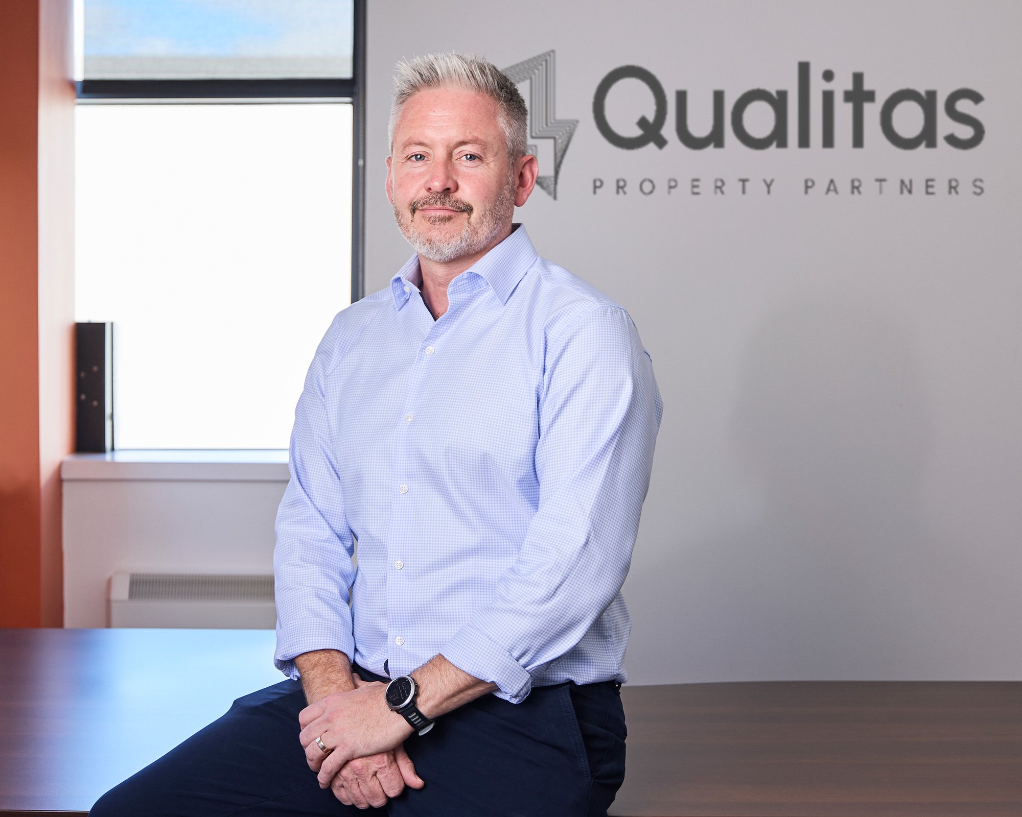 A middle-aged man with gray hair and a beard, wearing a light blue dress shirt, sitting on the edge of a table in an office. Behind him is a wall with the logo and name 'Qualitas Property Partners'.