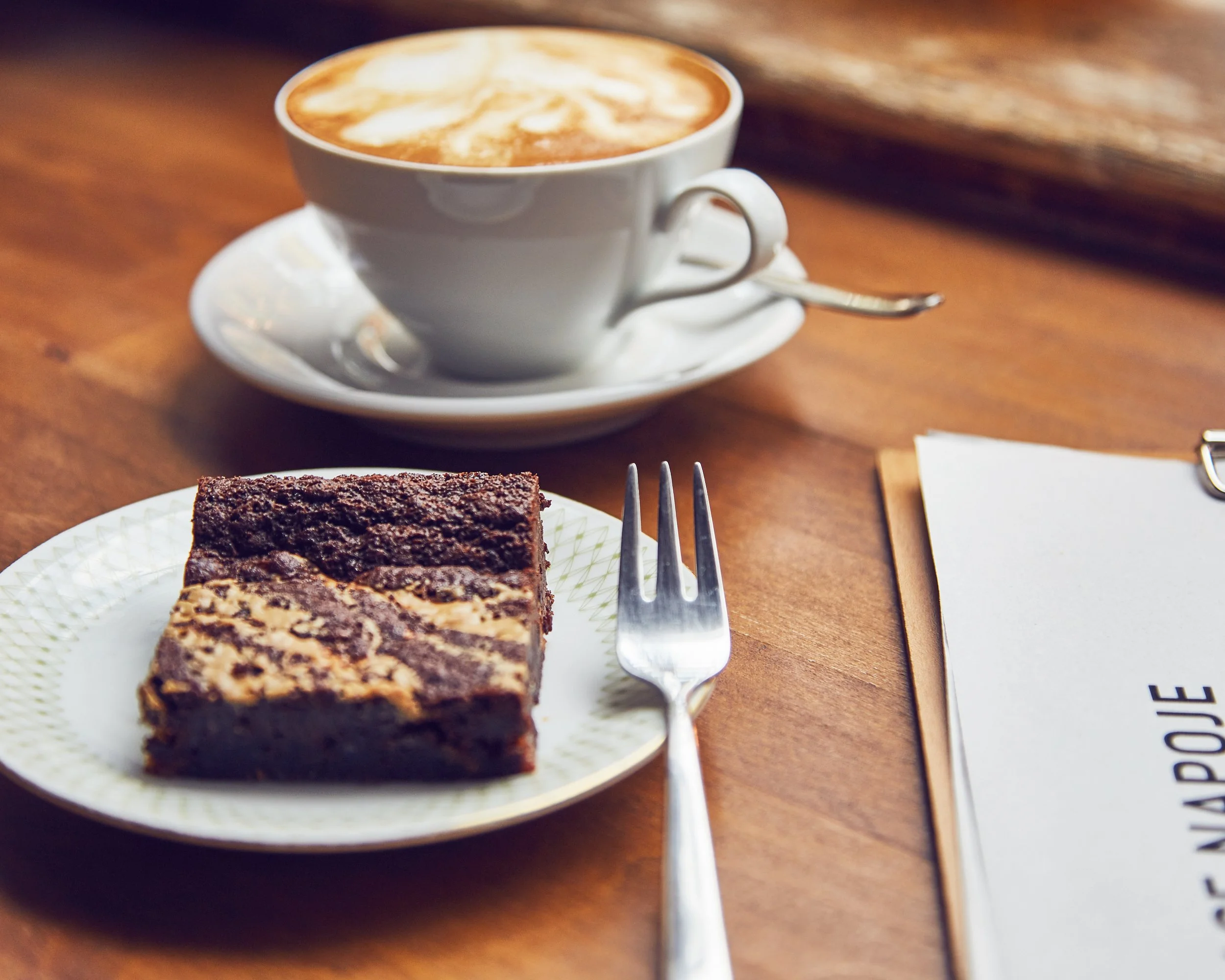 High end advertising profession product photo taken in location of a slice of chocolate marble cake on a decorative white plate, a fork nearby, a cup of coffee on a saucer, and a menu or notepad on a wooden table.