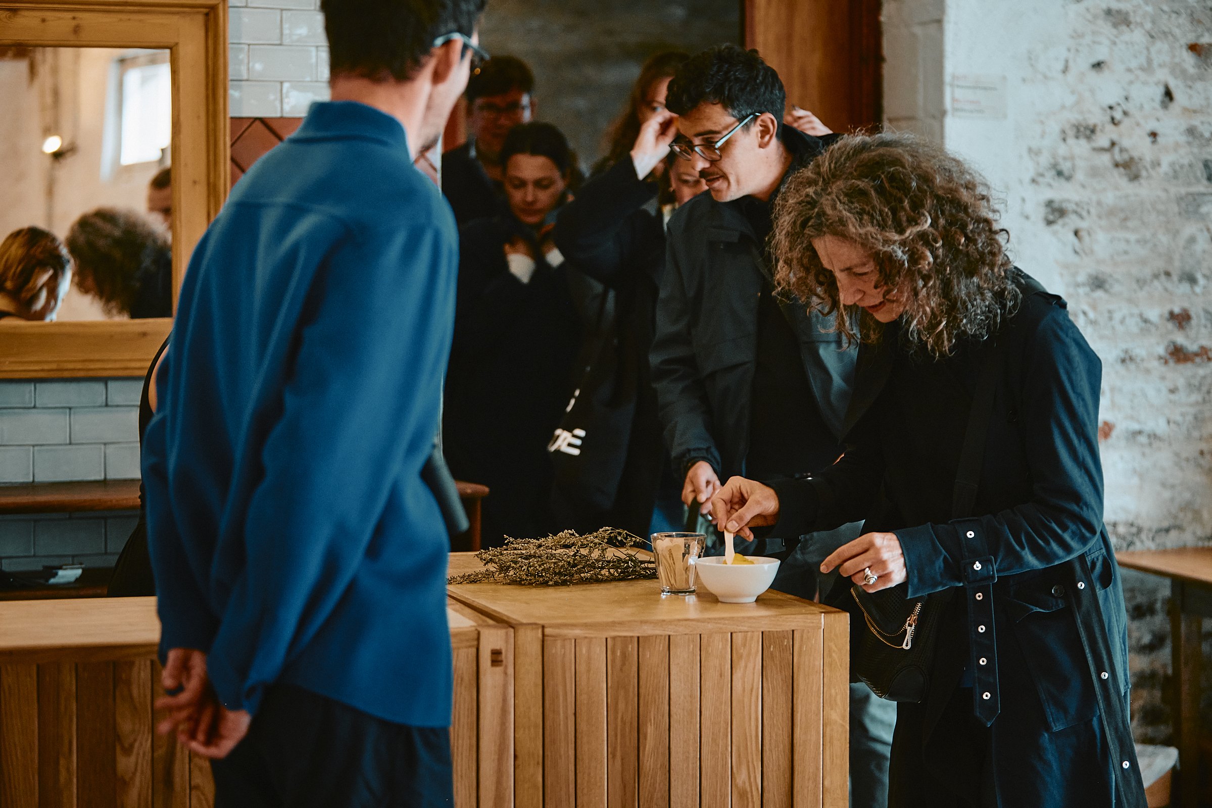 A group of people gathered around a wooden table indoors, some eating from bowls, with a woman serving food. The setting has a rustic vibe with exposed brick wall and wooden decor.