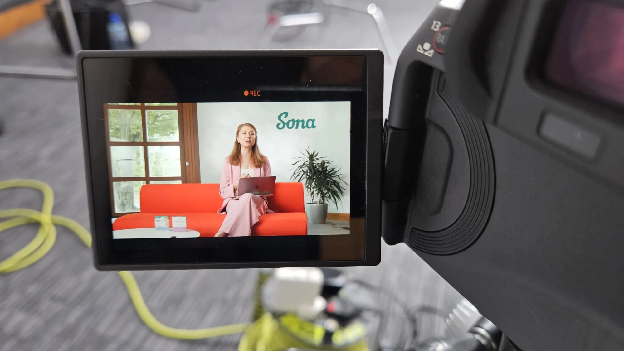 On a video camera display, a woman in a pink suit is sitting on a bright orange couch, holding a laptop and a phone, with a large window, green plants, and the name 'Sona' on a white wall behind her.
