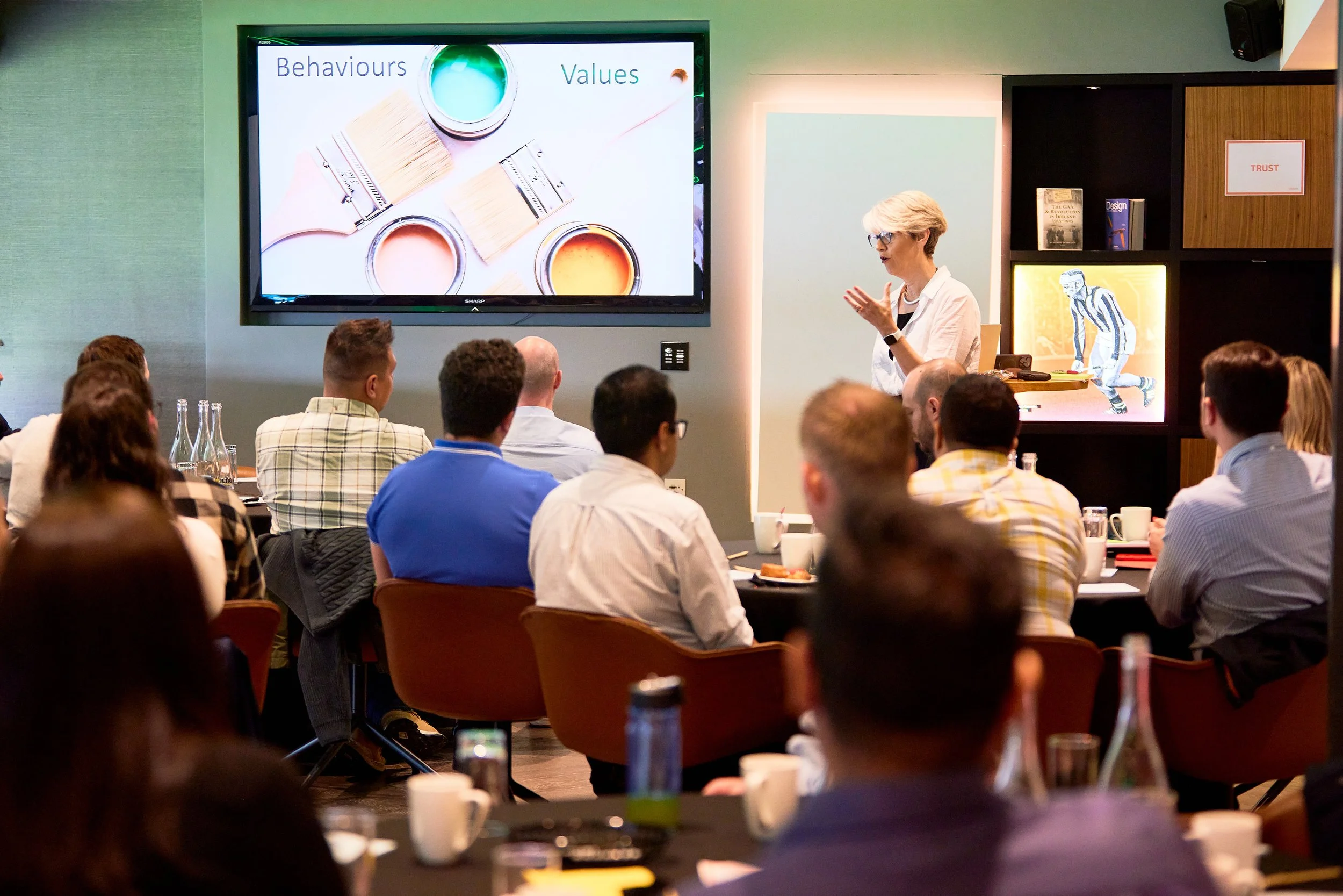 A woman giving a presentation in a conference room with a slide that says 'Behaviours' and 'Values' and shows paintbrushes and paint cans. Audience members are seated at tables, listening attentively.