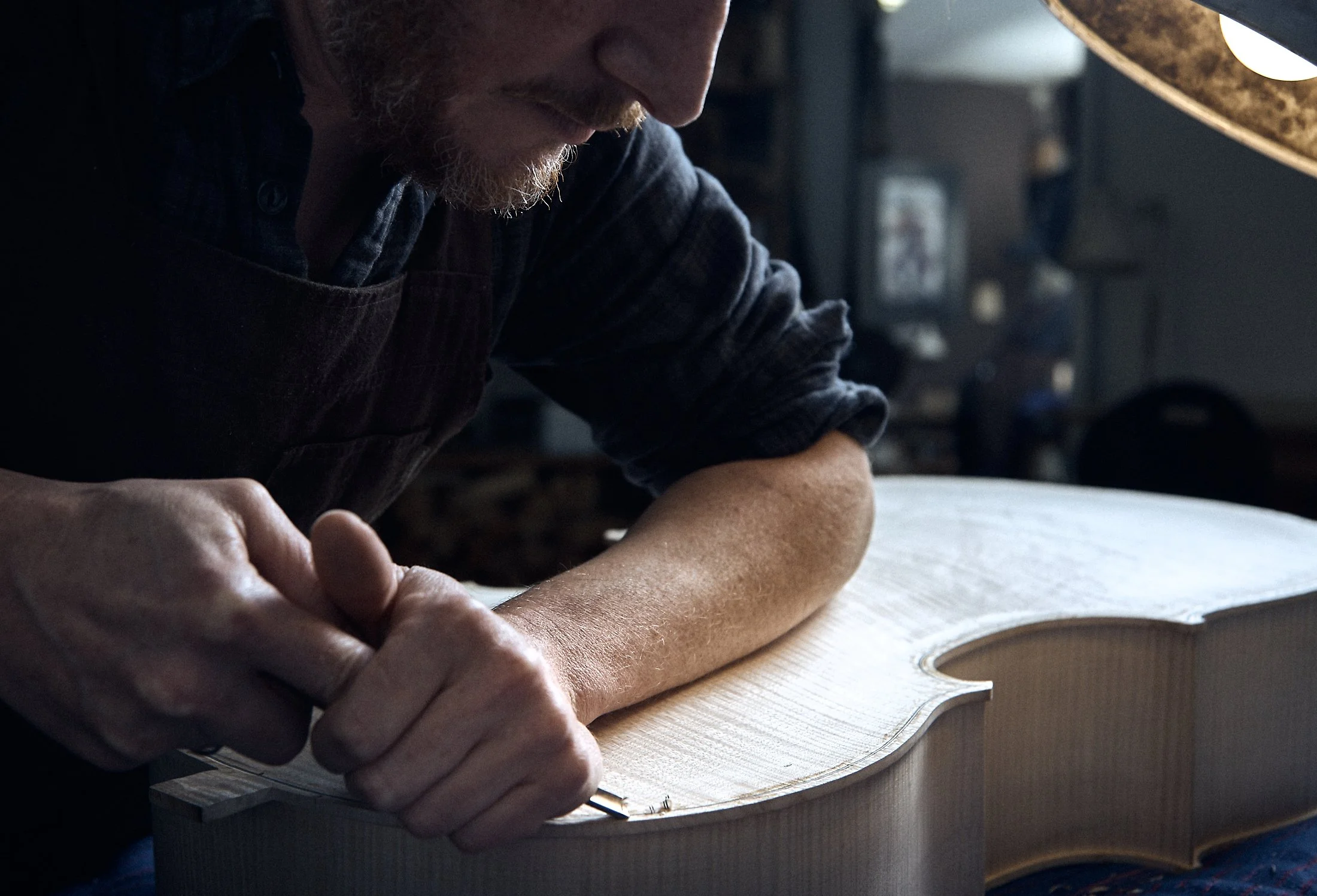 A man in a dark shirt and apron carefully works on a wooden guitar body, using a small tool, in a workshop. The workspace is dimly lit with a warm light.