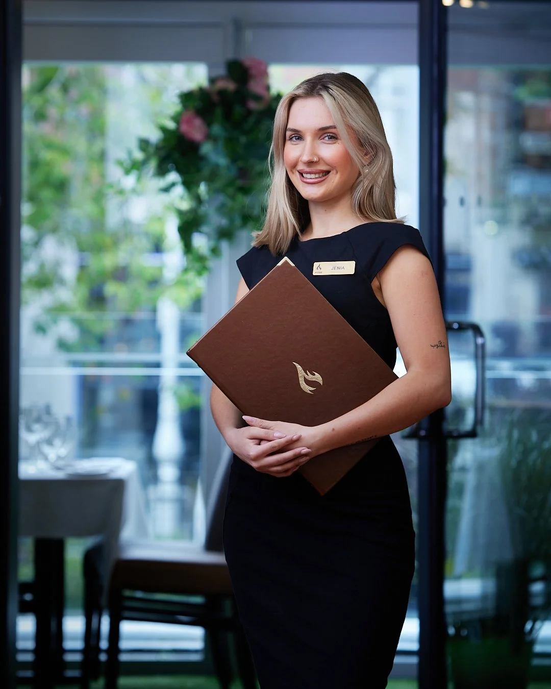 A young woman smiling and holding a menu inside a restaurant or cafe, wearing a black uniform with a name tag.