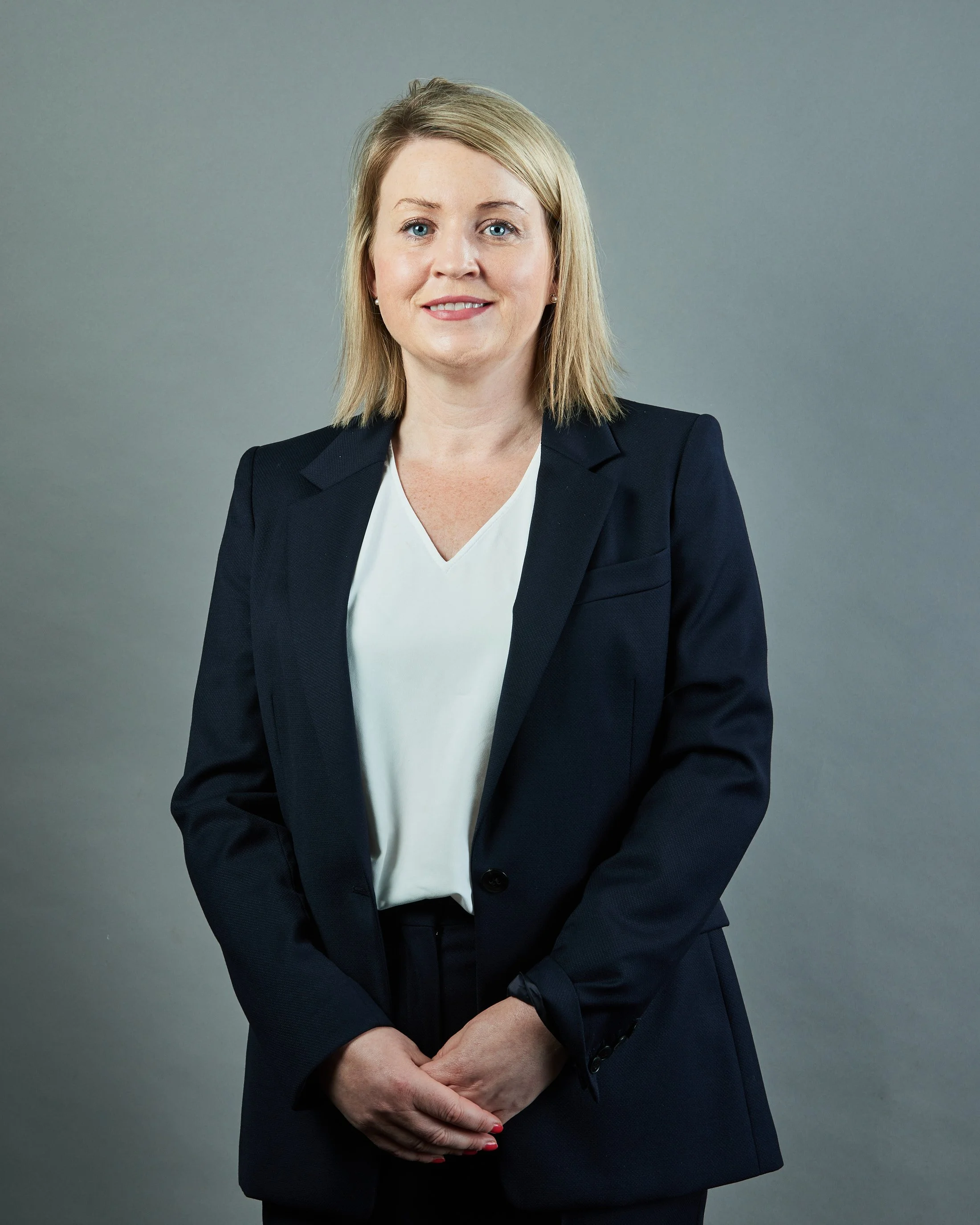 Professional corporate headshot of a CEO woman with blonde hair wearing a dark navy business suit and white blouse, standing against a plain gray background with profesional high quality photography lighting.