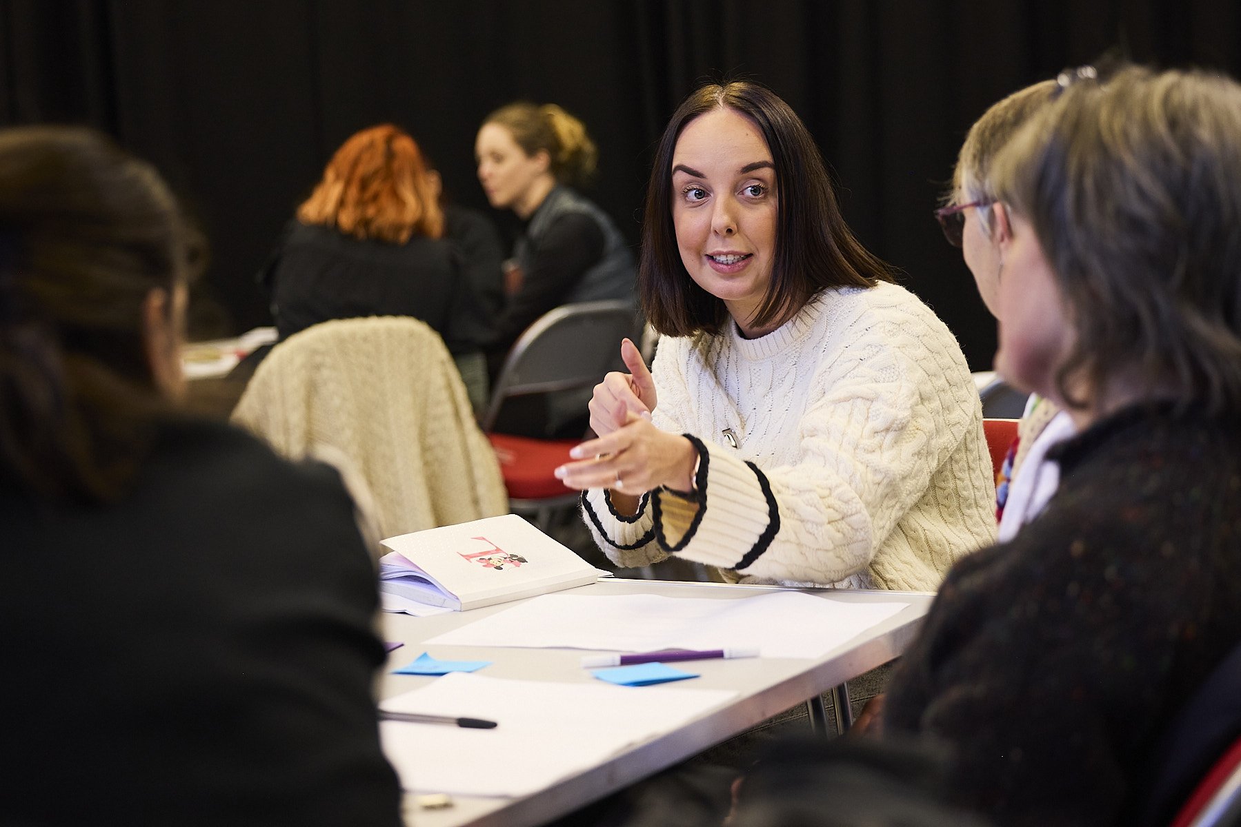 A woman with shoulder-length dark hair in a white sweater speaks to a group of women seated at a table during a discussion or meeting.
