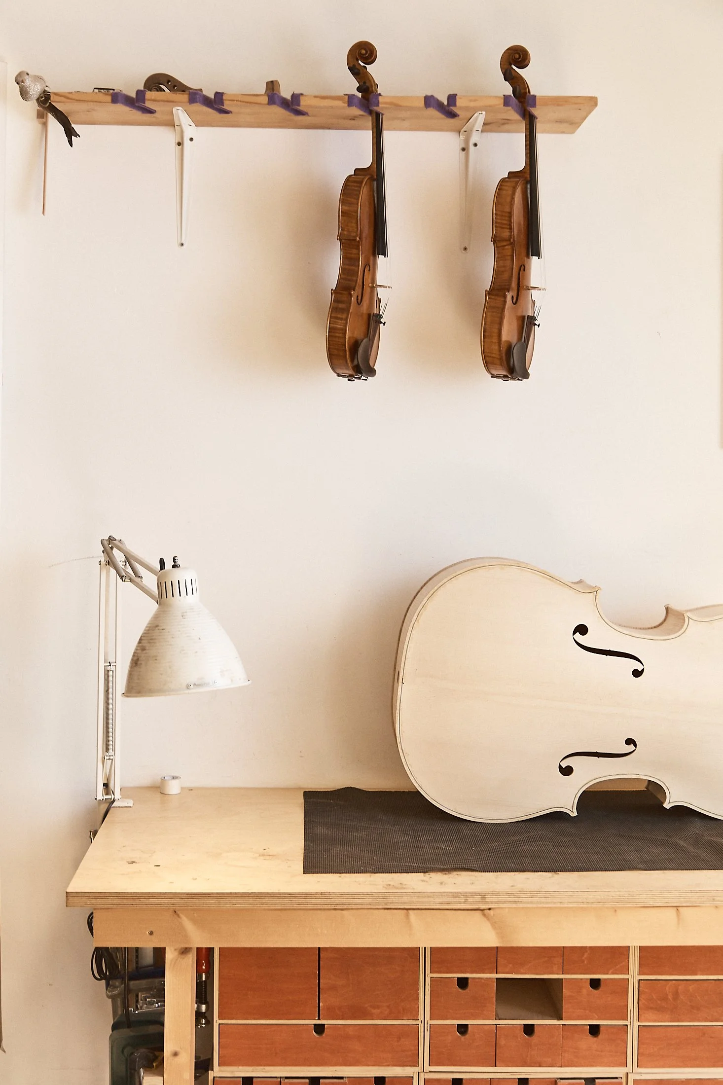 Two violins hanging on a wall above a workbench with a detached violin body and a desk lamp.