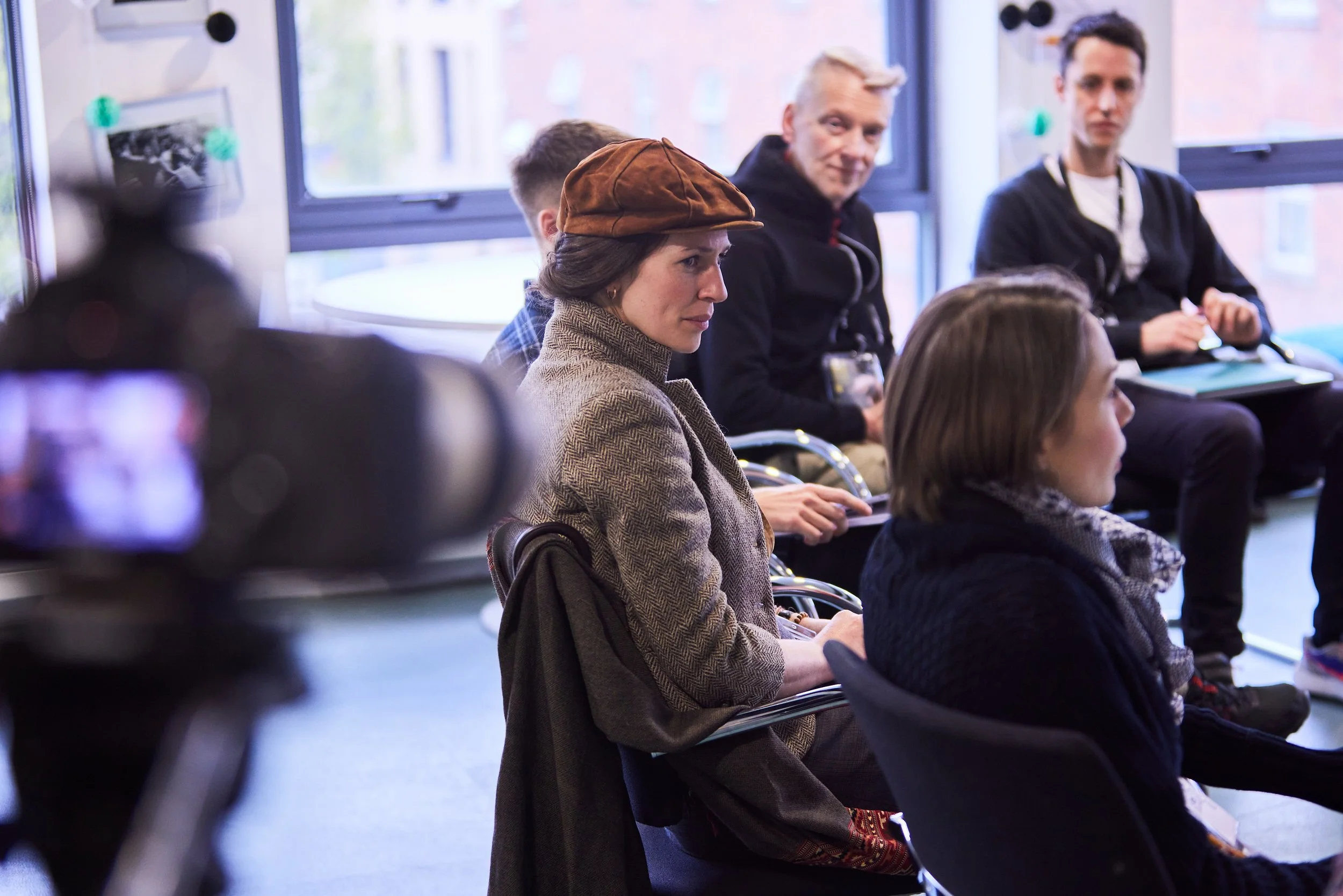 A woman with a brown hat and herringbone blazer sits in a conference room, surrounded by other seated people, with a camera visible in the foreground and windows behind.