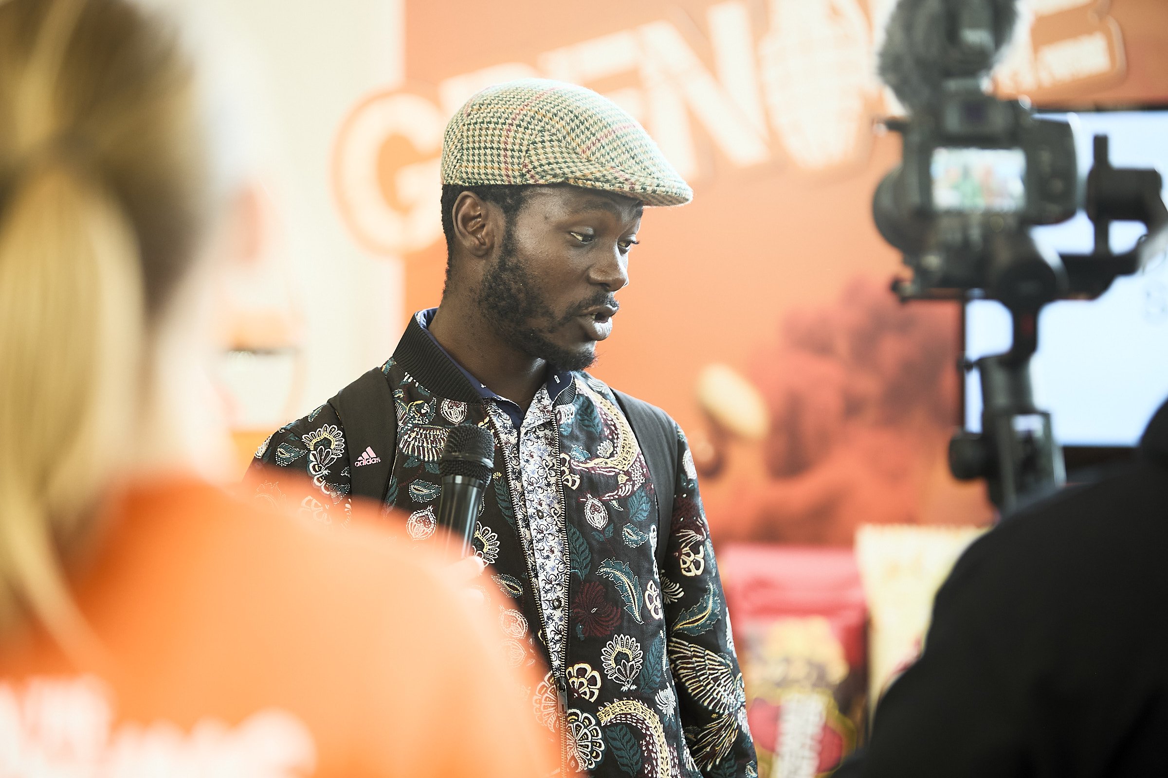 A man wearing a patterned blazer and a flat cap speaking into a microphone during an interview, with a camera filming him and a blurred person in the foreground.