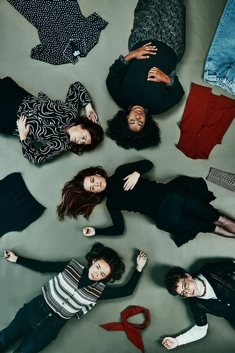 Promotional image of five women lying on the floor surrounded by various pieces of clothing, with three of them looking at the camera, and two with eyes closed or looking away, shooting in location  with professional photography lighting.