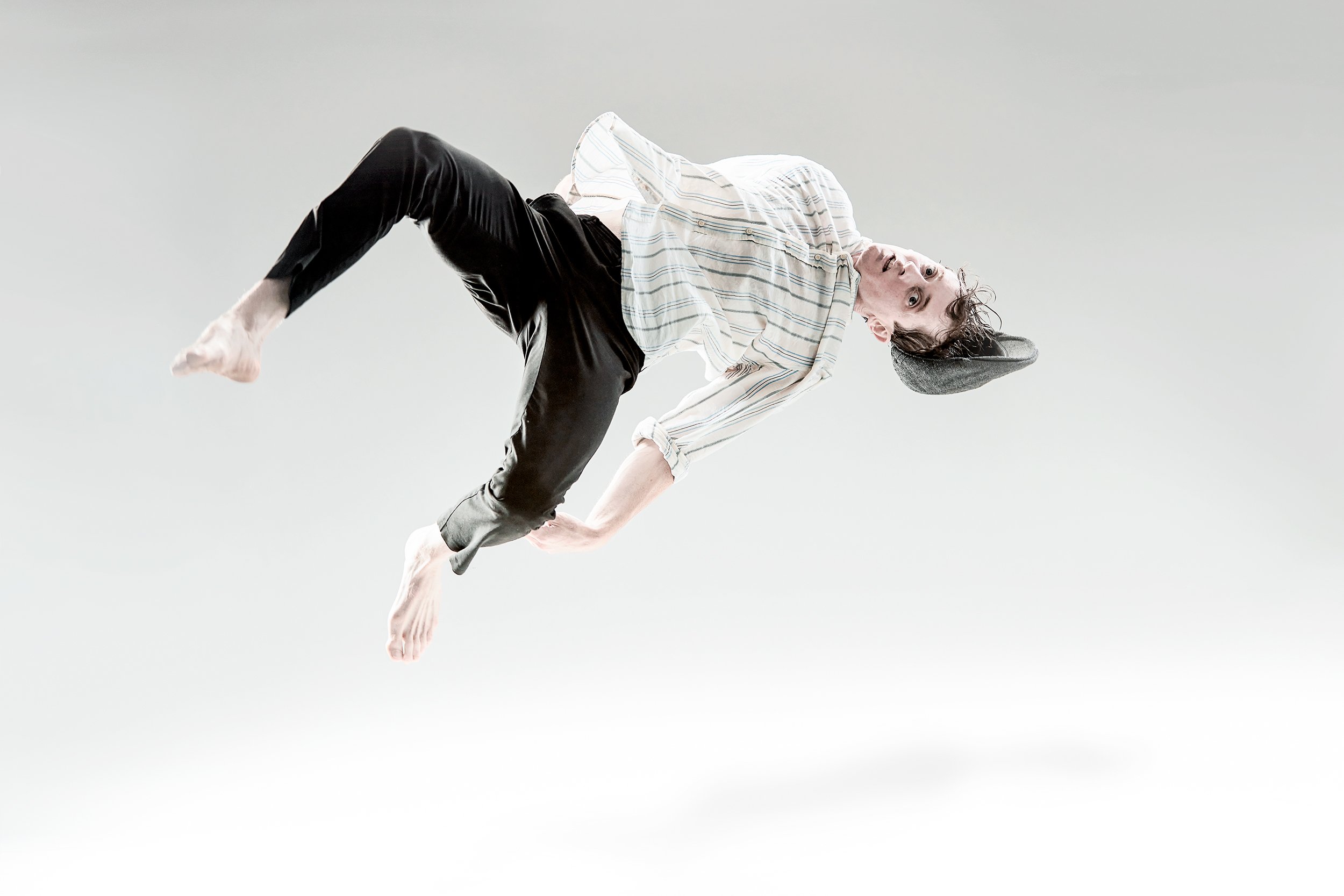 Strong commercial photo of a young man dressed in a striped shirt, black pants, and a gray beanie, floating in mid-air against a plain light background with an expression of surprise or curiosity, in a photo studio with professional lighting.