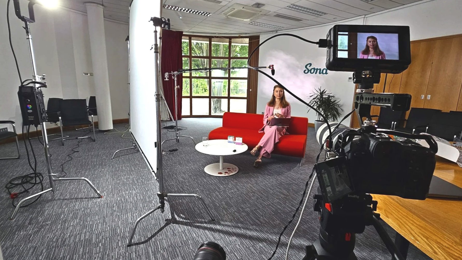 A woman in pink attire sitting on a red sofa during a video recording session, with filming equipment and a large window with red curtains in the background.