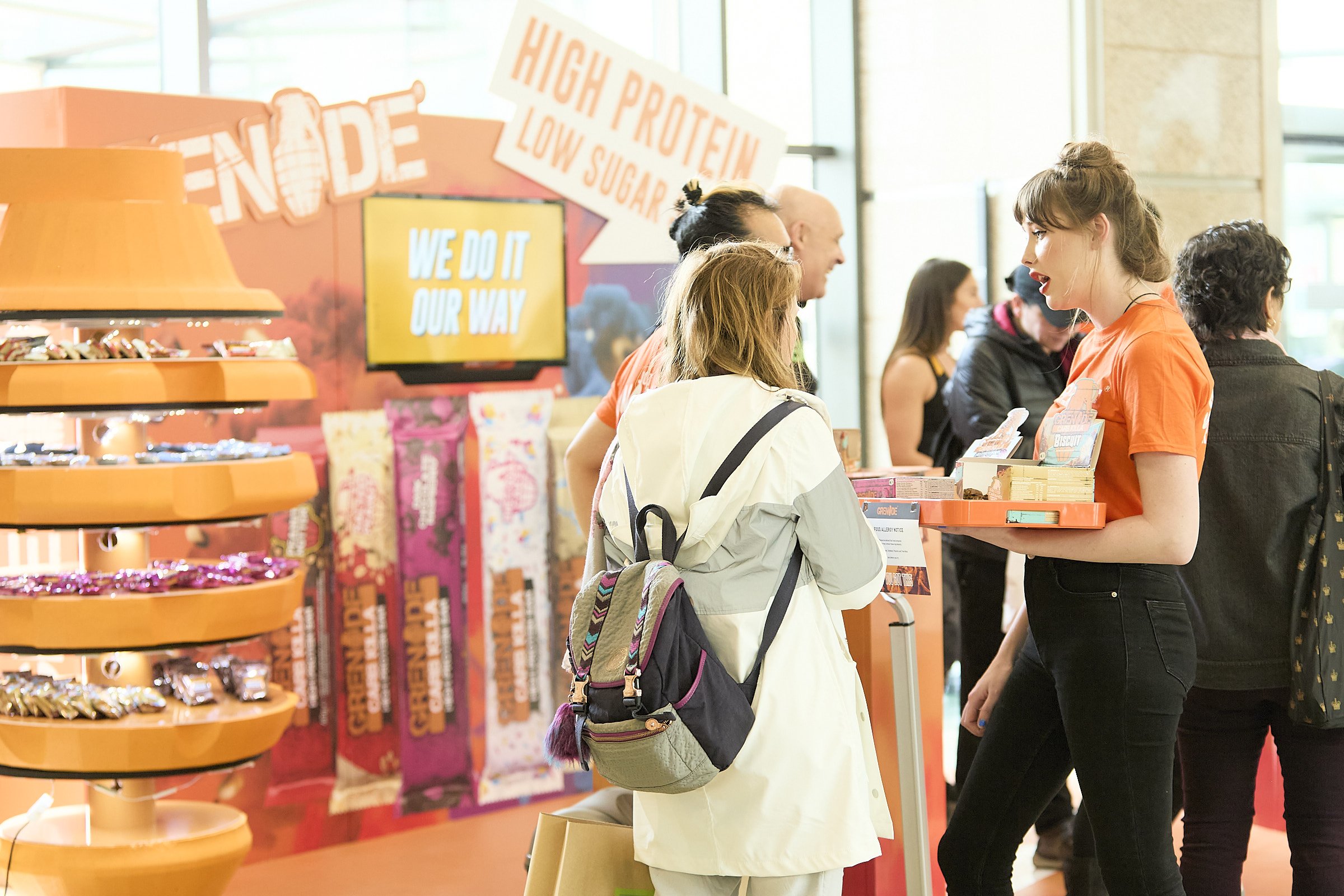 A young woman in an orange shirt is talking to a customer at a booth in a shopping mall. The booth has bright signage promoting high-protein, low-sugar snacks. The customer is wearing a white jacket and has a backpack. Other shoppers are visible in t