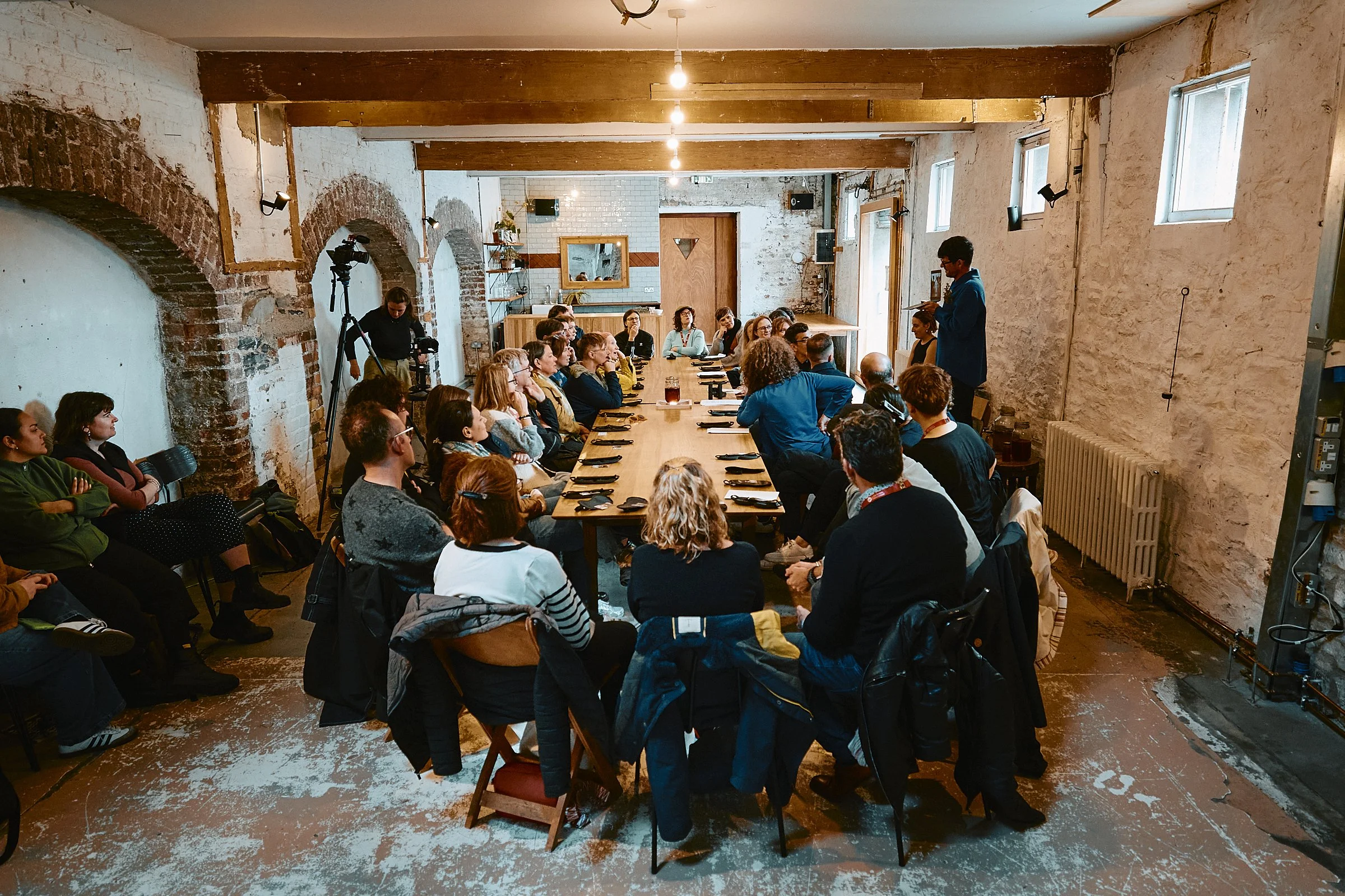 Group of people attending a presentation or meeting in a rustic room with exposed brick walls and wooden beams, seated around a long wooden table.