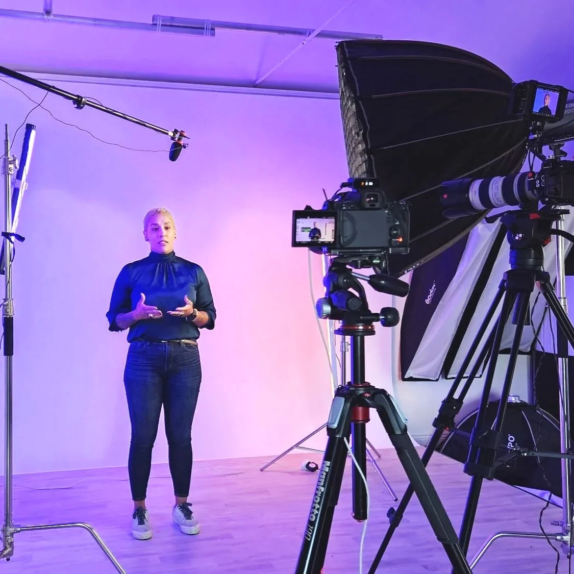 A woman is speaking or presenting in a photography studio with professional lighting and filming equipment set up around her.