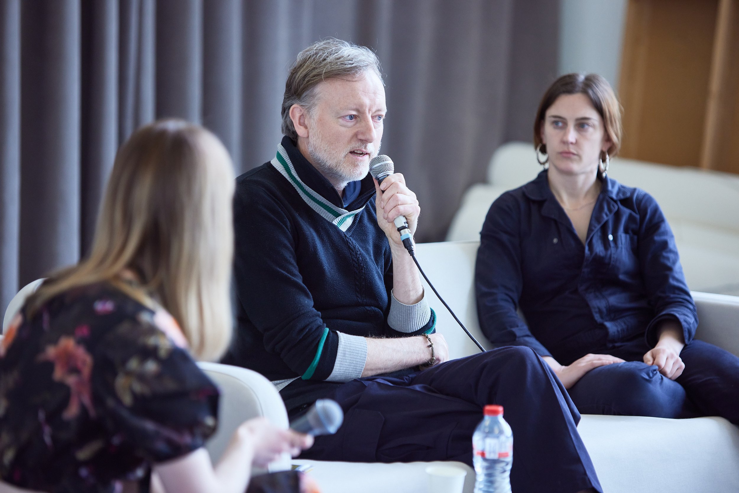 A panel discussion with three people seated on a sofa, one man speaking into a microphone, a woman holding a microphone, and another woman listening.