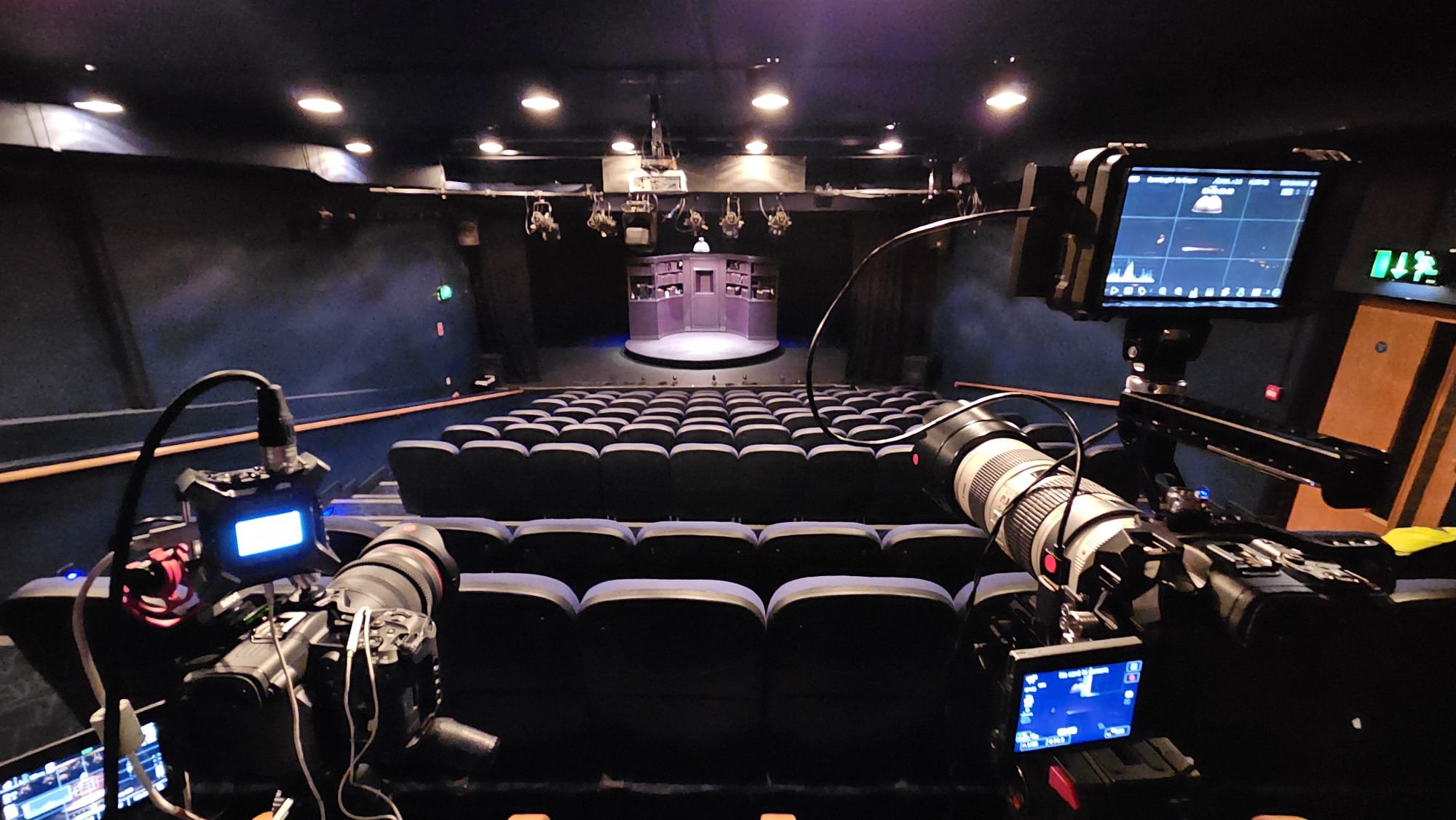 Interior of a theater or performance space with empty seats facing a stage with purple curtains. Filming or photography equipment is set up in the foreground, including cameras and monitors.