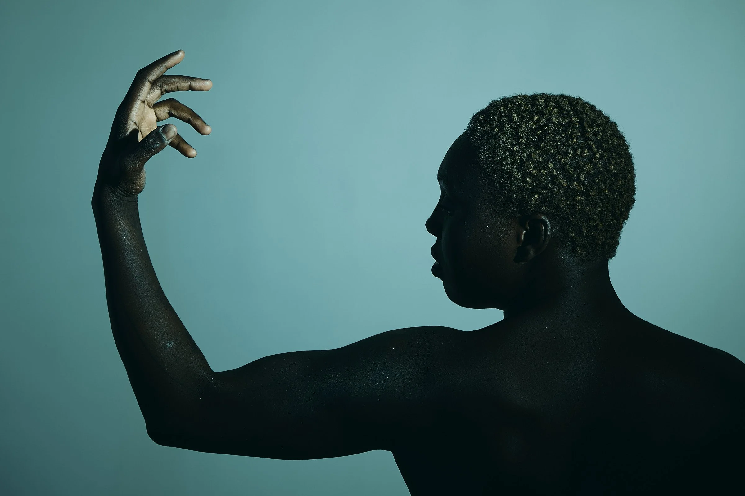 A silhouette of a person with short, curly hair, posing with their right arm bent and hand near their face against a plain teal background.